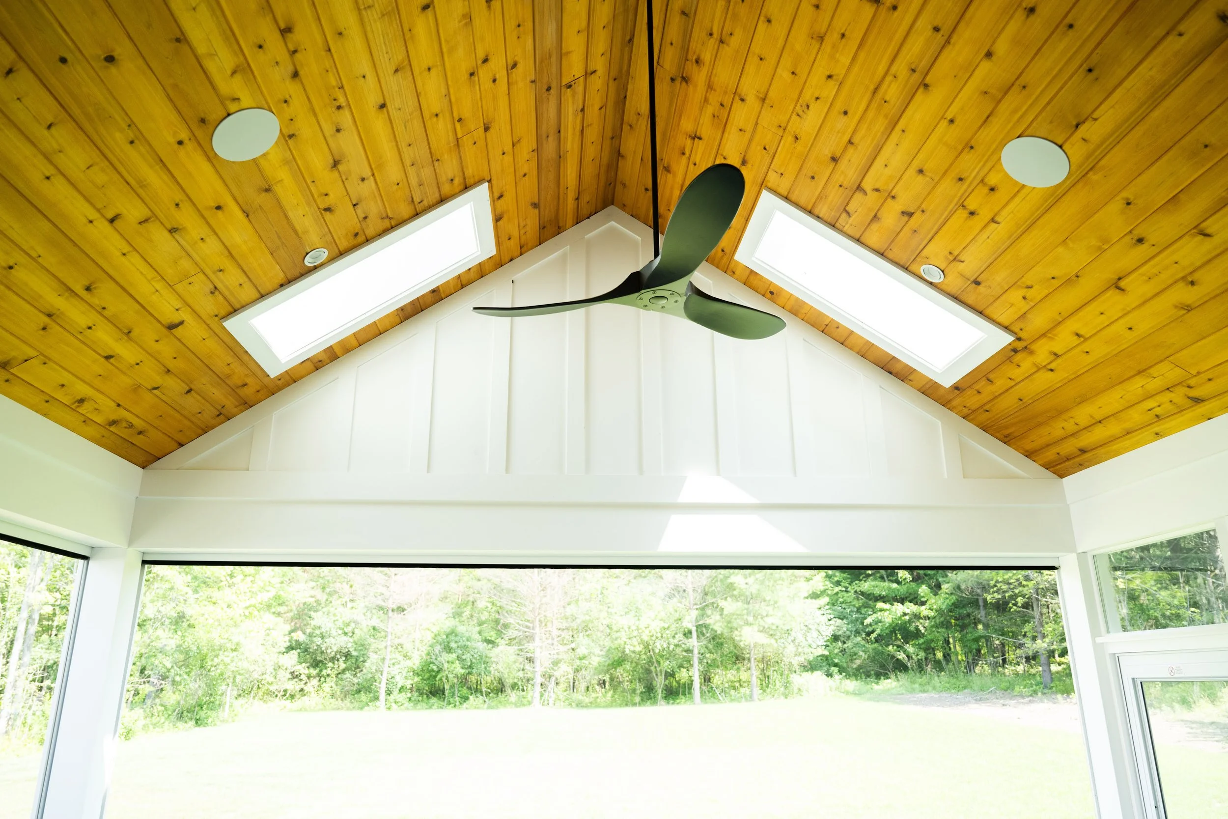Ceiling with wooden planks, two skylights, and a ceiling fan, overlooking windows and a lush green outdoor landscape.