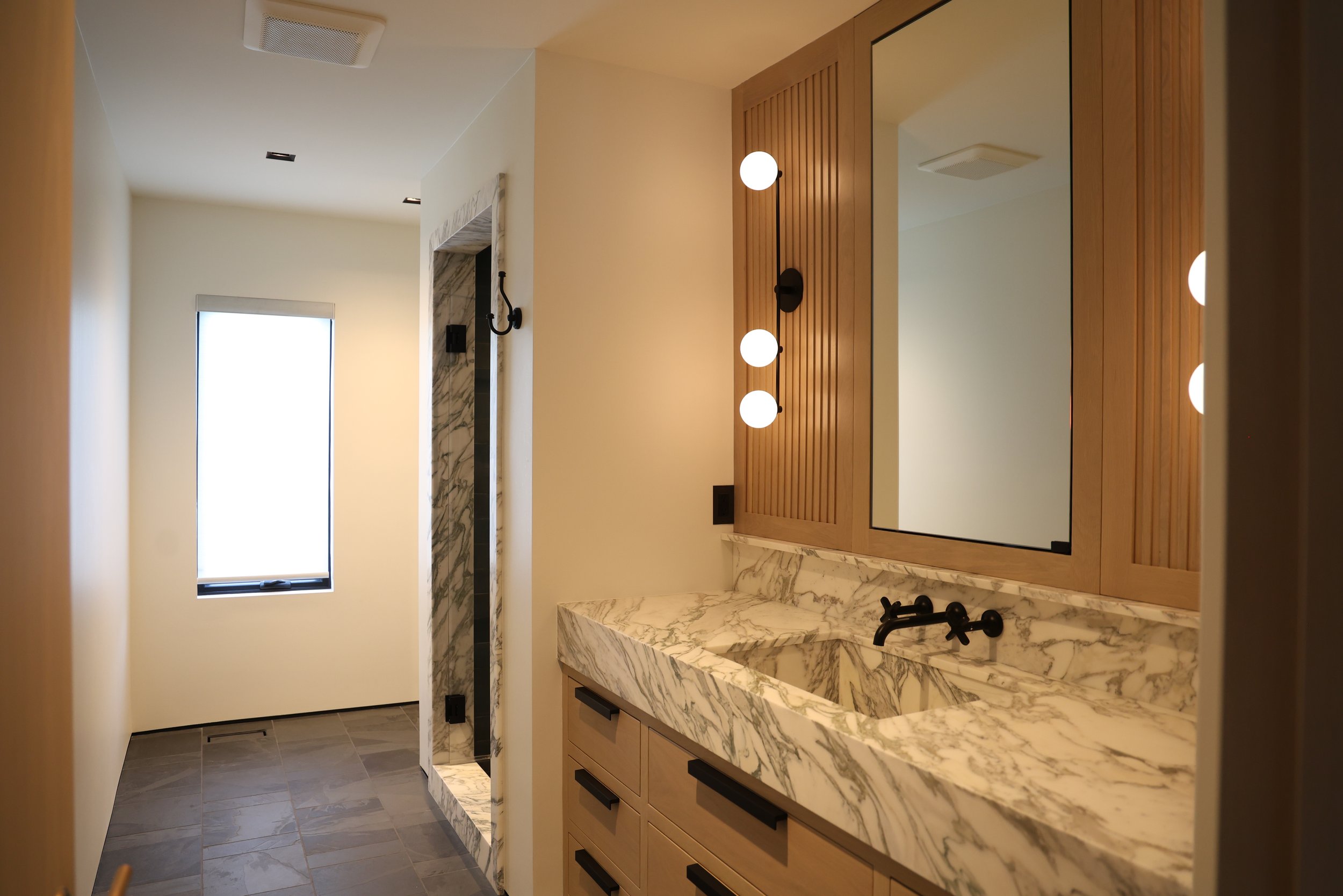 Modern bathroom with marble countertop, wooden cabinet, round mirror, black faucet, black light fixtures, and a window letting in natural light.