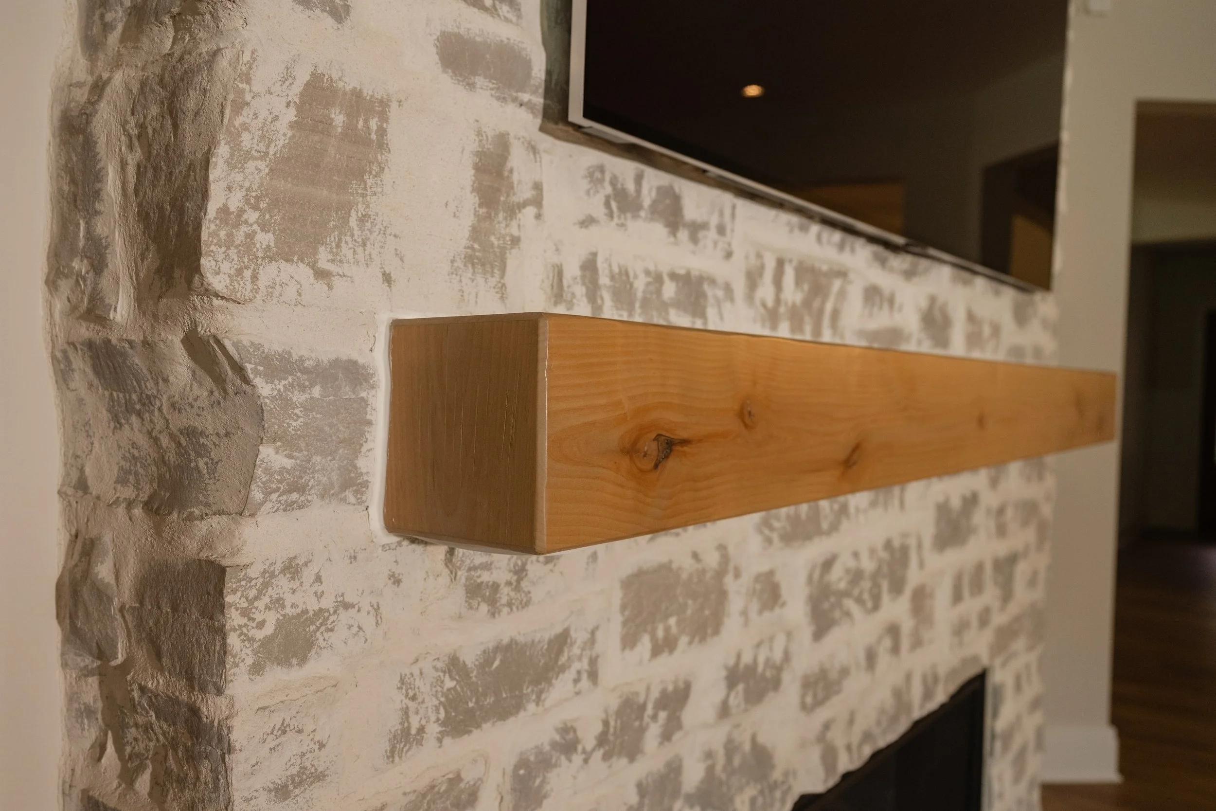Close-up of a textured brick wall painted white, with a horizontal strip of wood at the bottom, and a flat-screen television mounted on the wall.