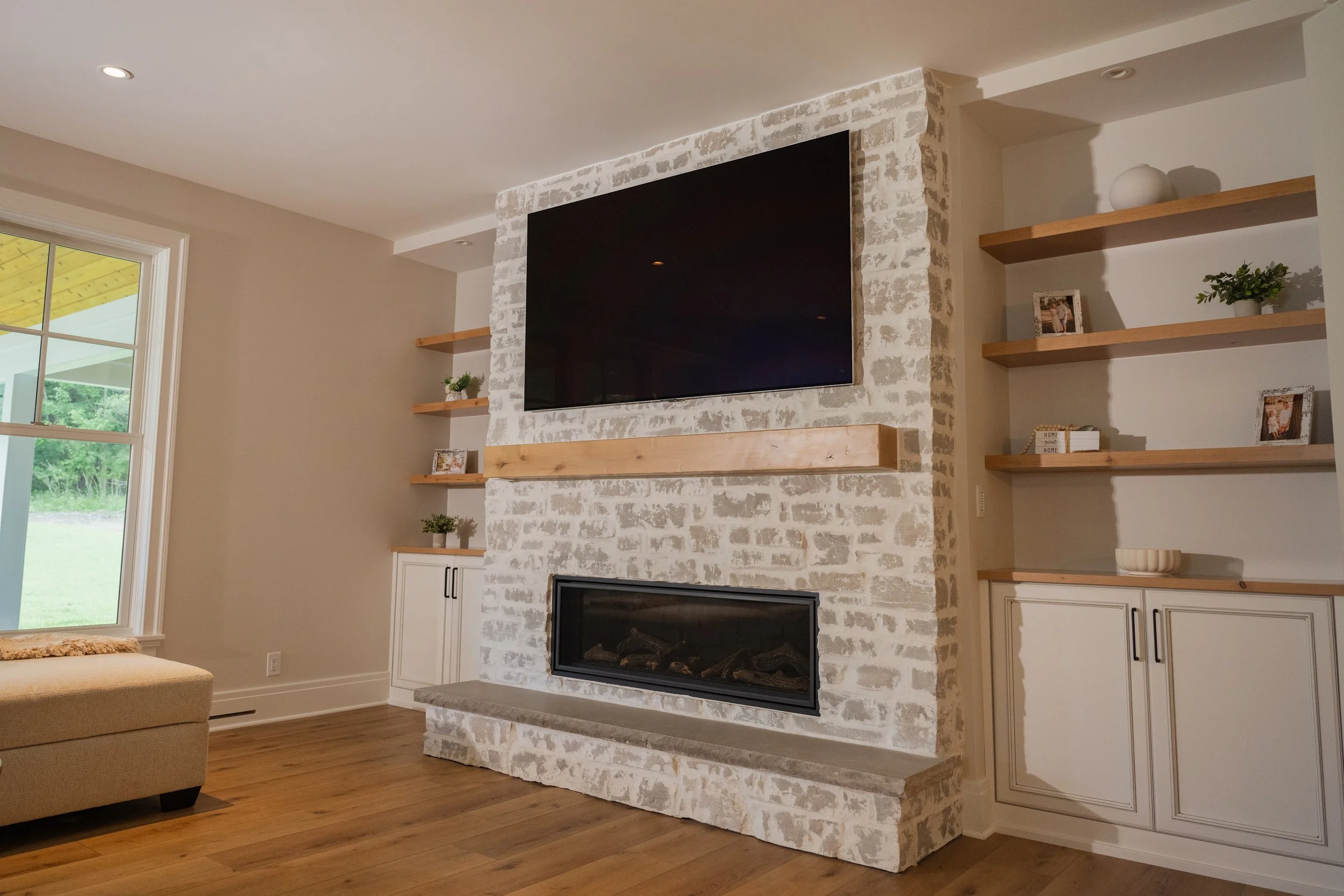 Living room with a white brick fireplace, a large flat-screen TV mounted above, wooden shelves with decorative items, a window showing a yard outside, and a beige couch in the foreground.