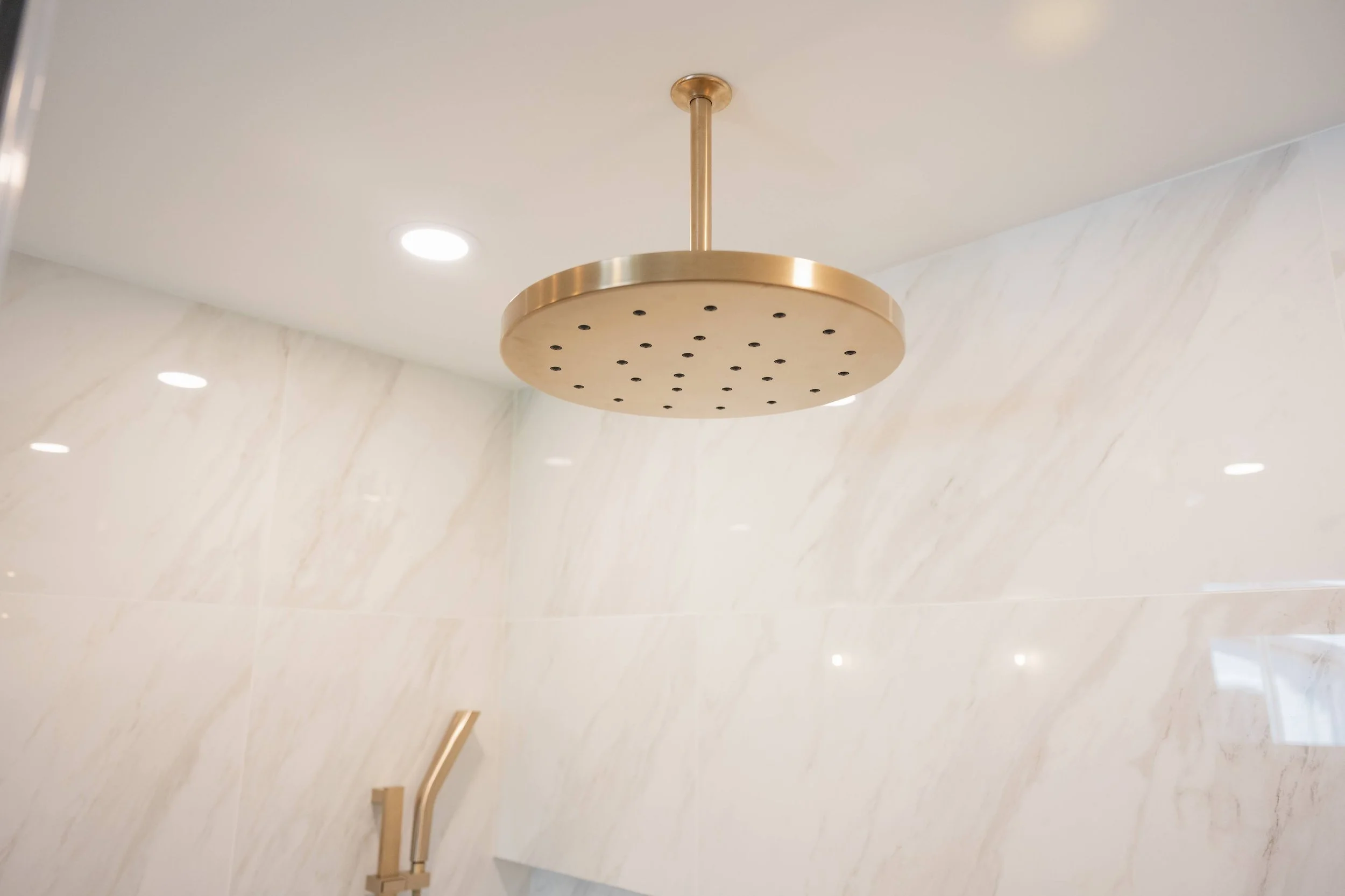 Photo of a round, gold-colored showerhead attached to a white ceiling in a bathroom with beige marble walls.