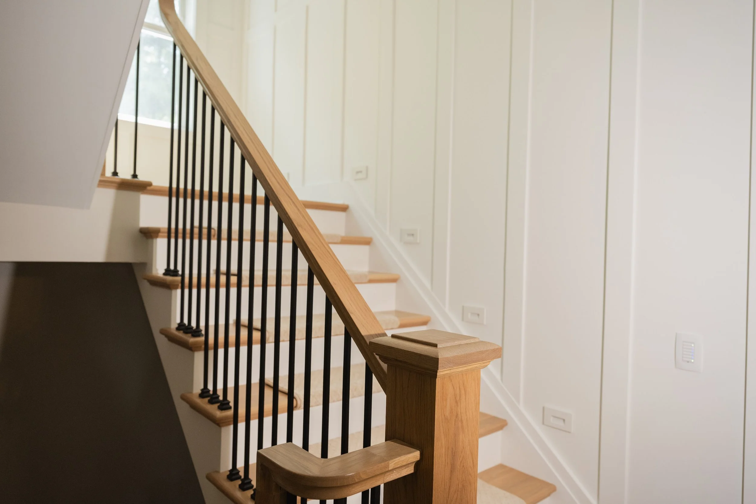 Interior view of a staircase with wooden steps, black metal balusters, and a wooden handrail, leading up beside a white-paneled wall and a window with natural light.