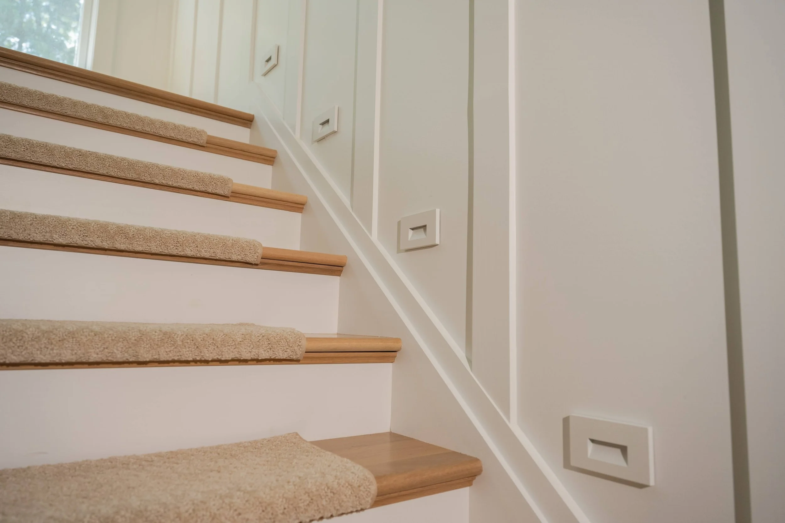 Interior view of a staircase with beige carpeted steps, white risers, and wood trim, next to a white wall with decorative square moldings and a window letting in natural light.