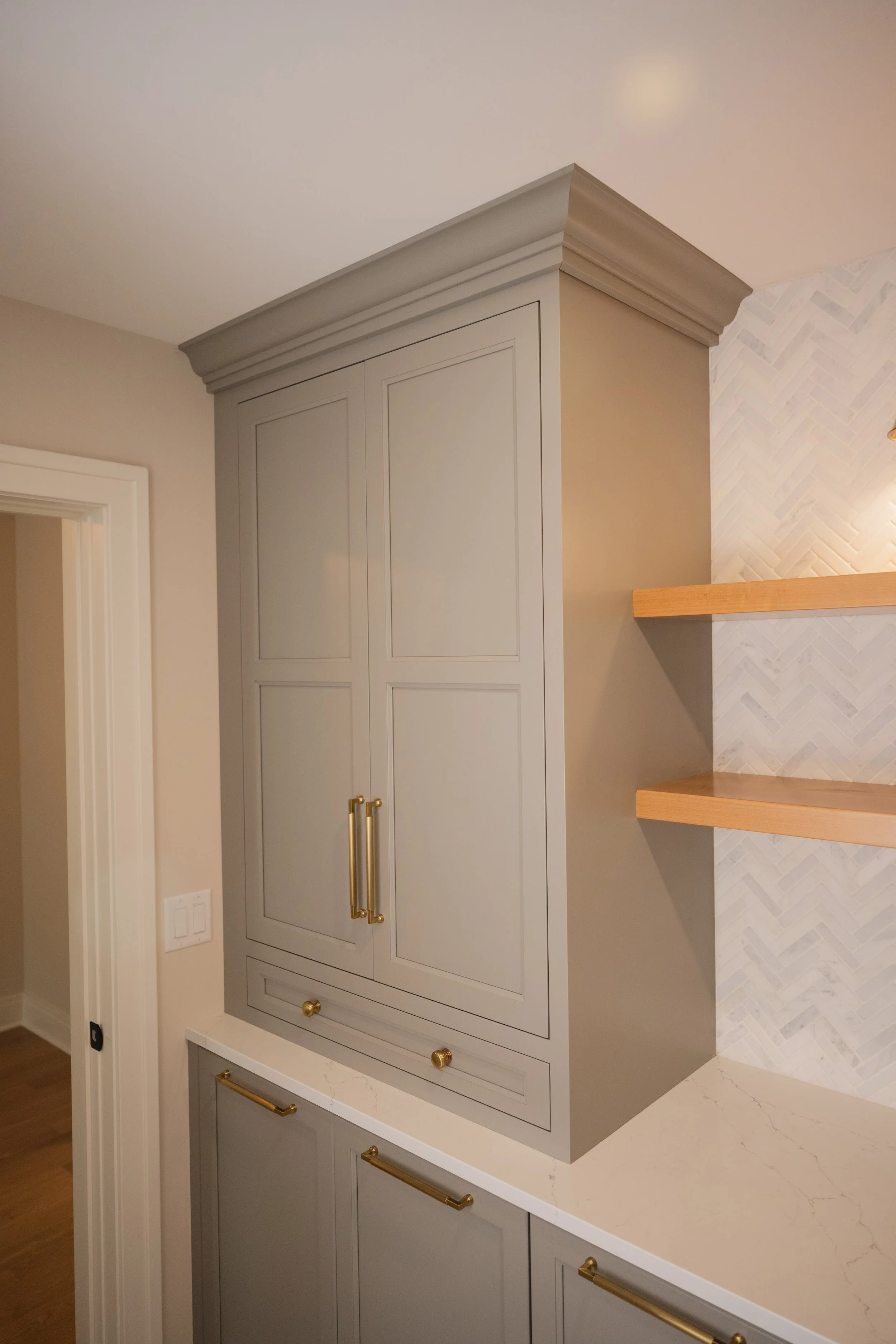 A kitchen corner with a built-in cabinet painted in light grey, featuring gold handles, against a wall with herringbone-patterned tile and a wooden shelf.
