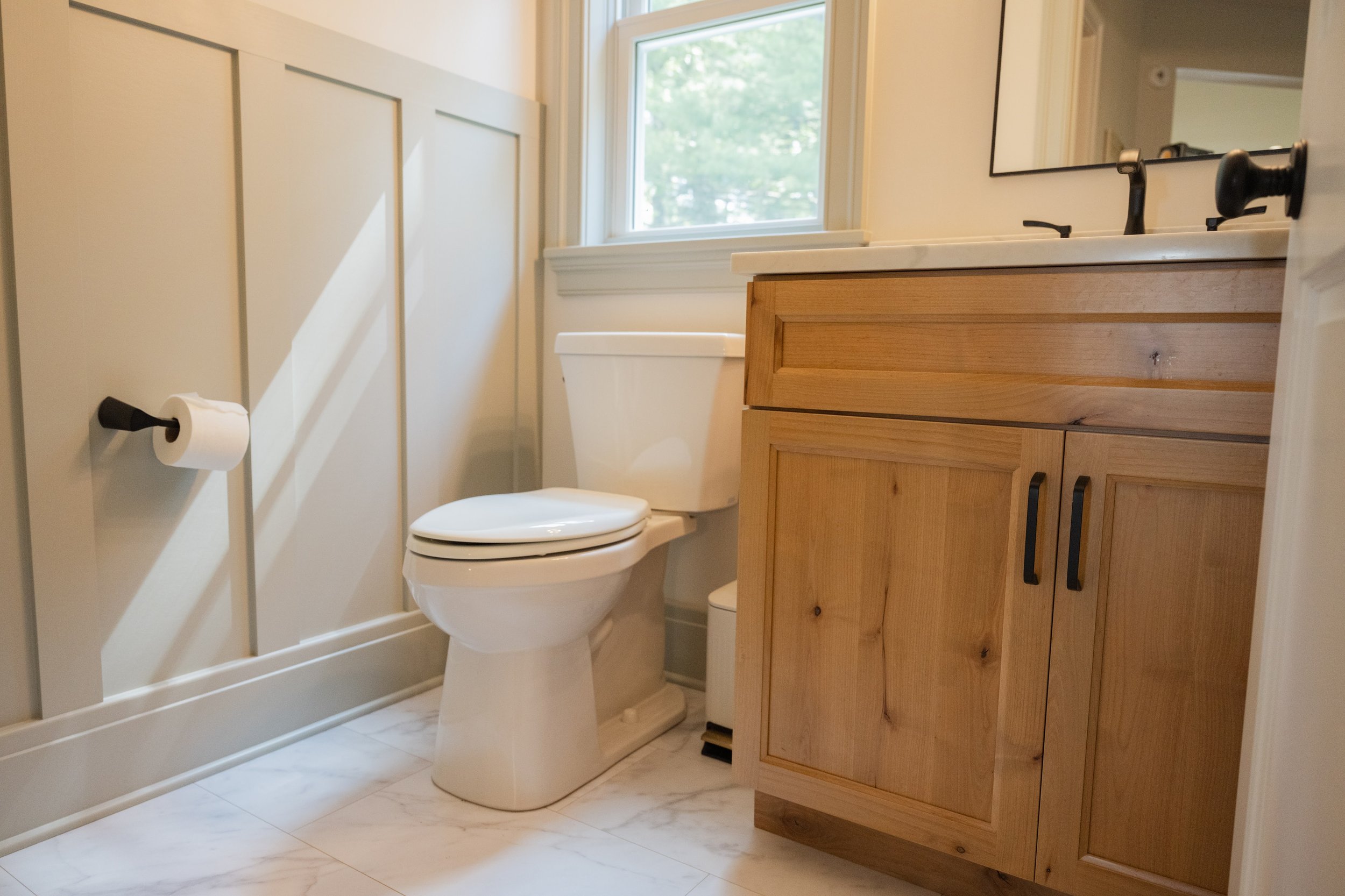 A small bathroom with a toilet and a wooden vanity with a white countertop and black hardware. A window above the toilet lets in natural light. The walls are painted in a light neutral color, and the floor is tiled with white marble-like tiles.