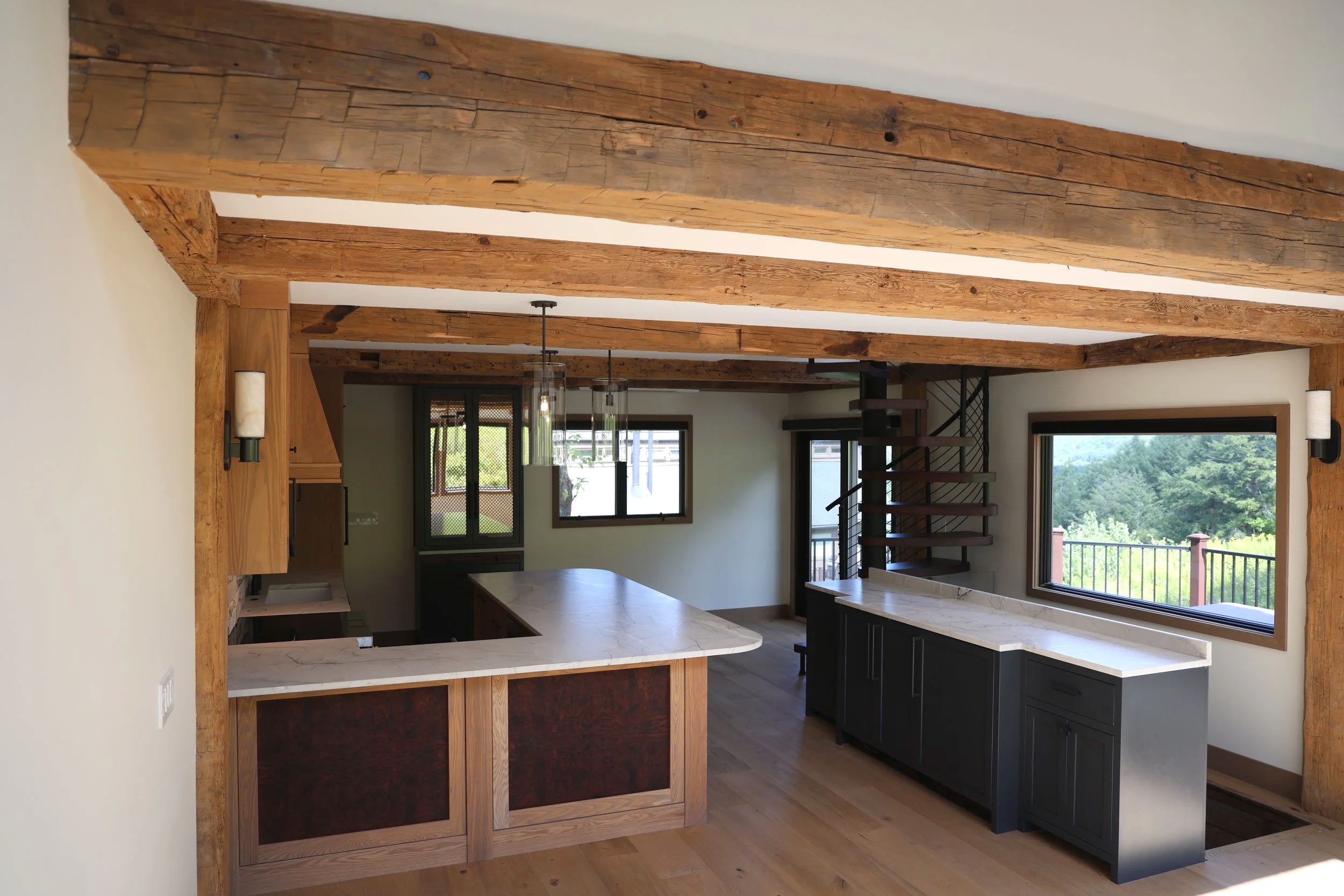 Interior view of a kitchen with exposed wooden beams on the ceiling, a kitchen island with a white countertop, black cabinetry, large windows showing a green outdoor landscape, and modern pendant lights hanging above the island.