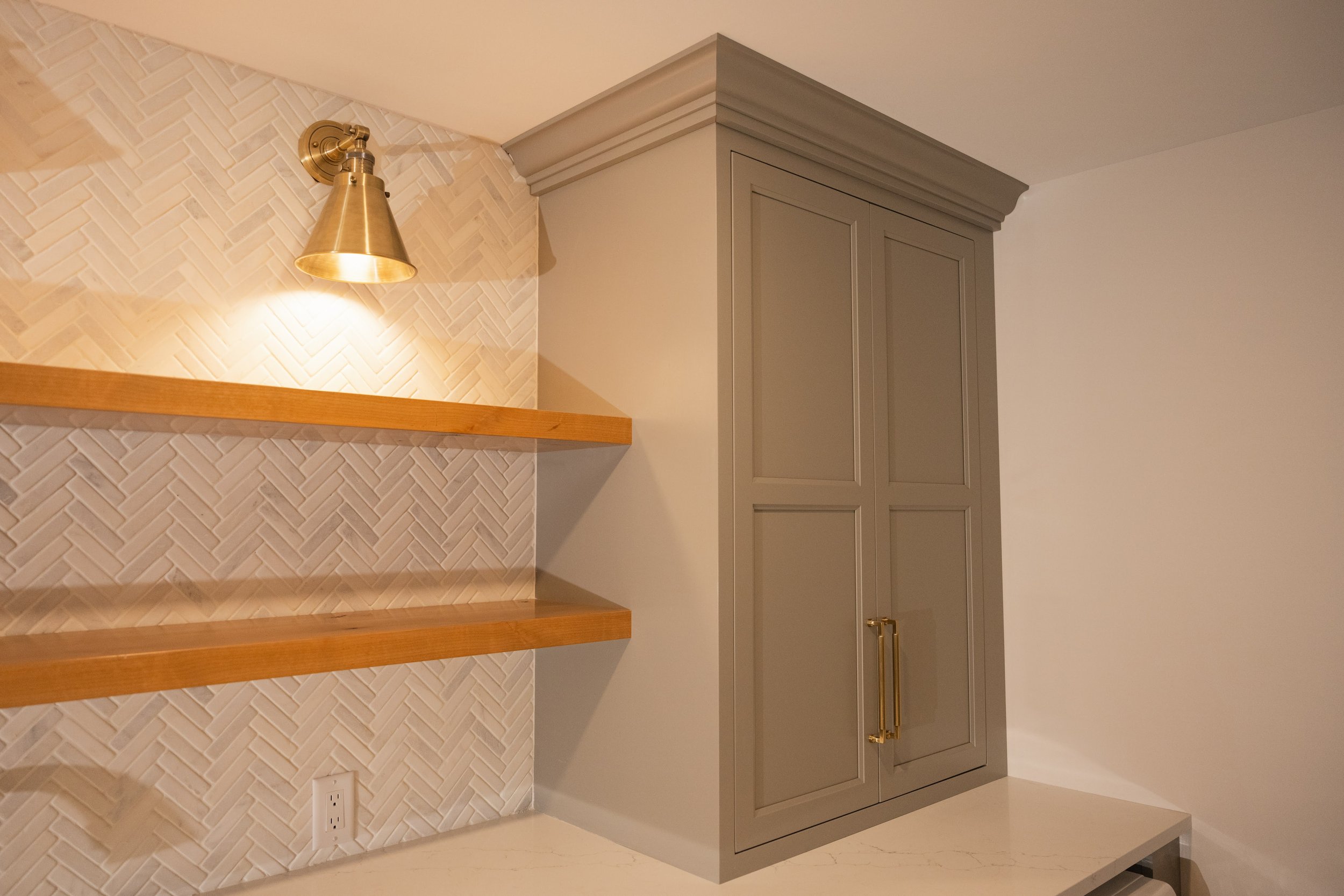 Interior kitchen corner with grey cabinetry, beige crown molding, decorative backsplash, gold-tone wall light, and electrical outlet.
