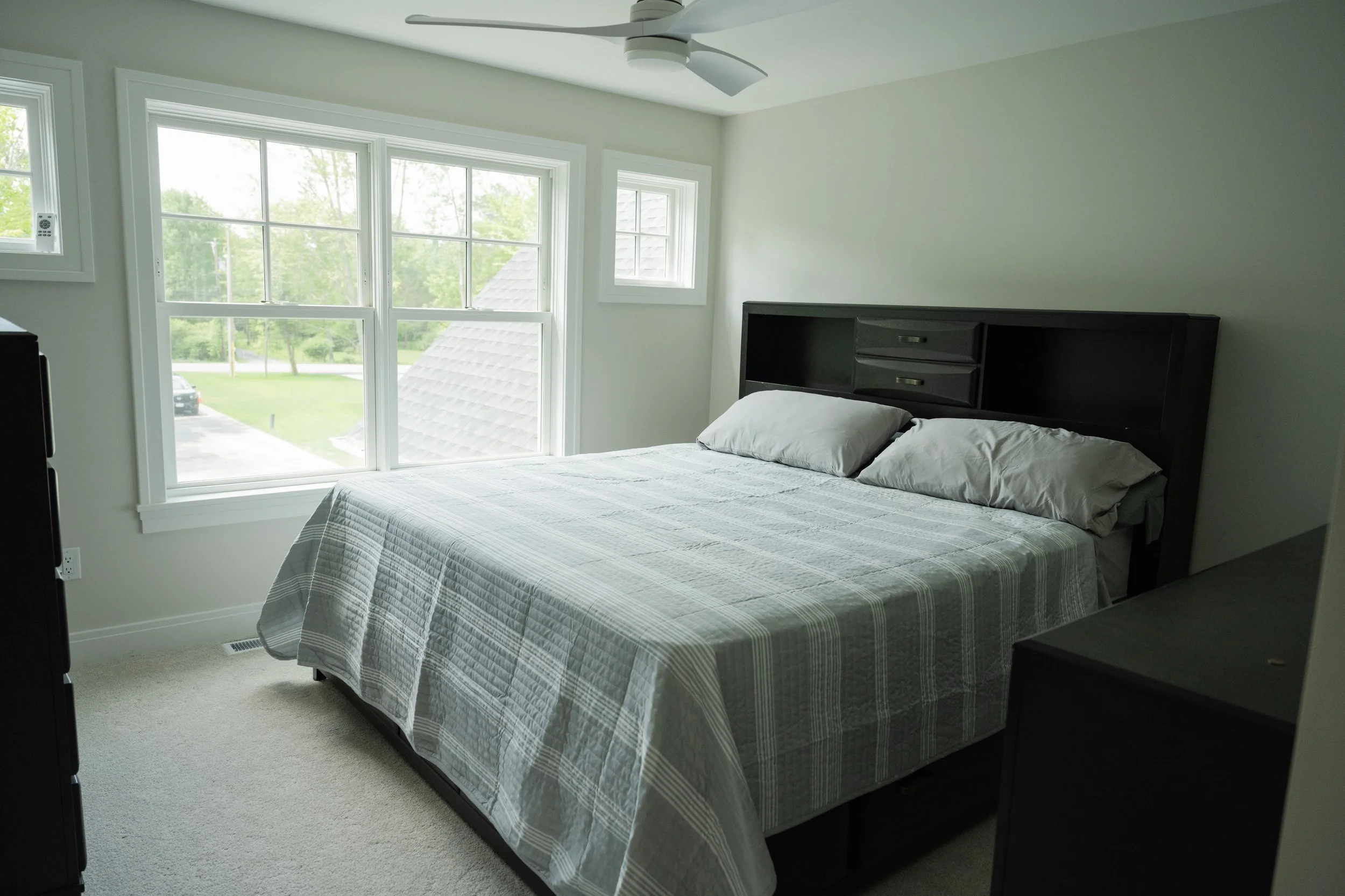 A bedroom with a bed, black headboard, white quilt, and two pillows, next to three large windows with white trim showing trees and a driveway outside, ceiling fan, and beige carpet.