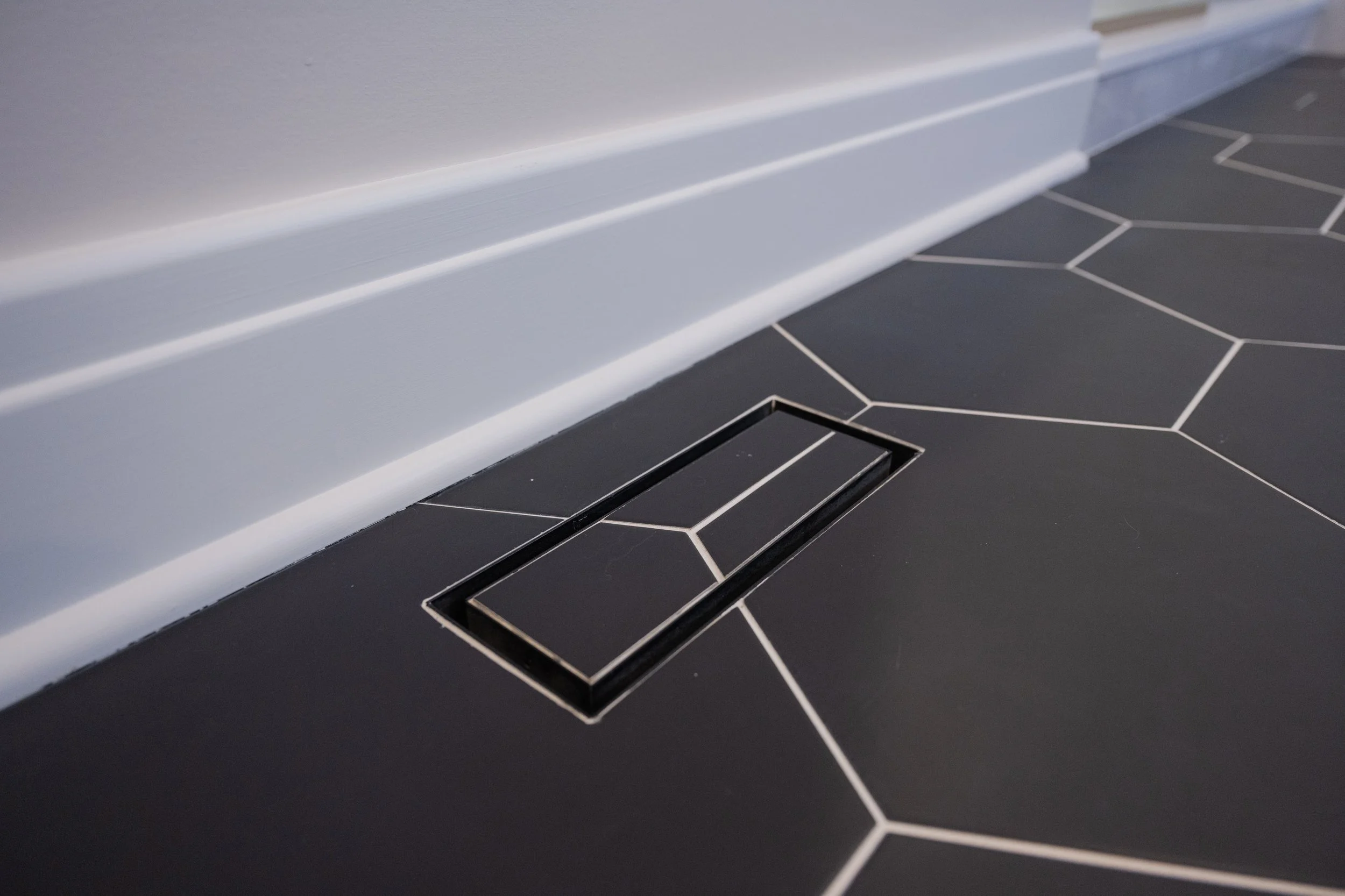 Close-up of a black tiled floor with white grout lines and a floor drain, along with a white baseboard along a gray wall.