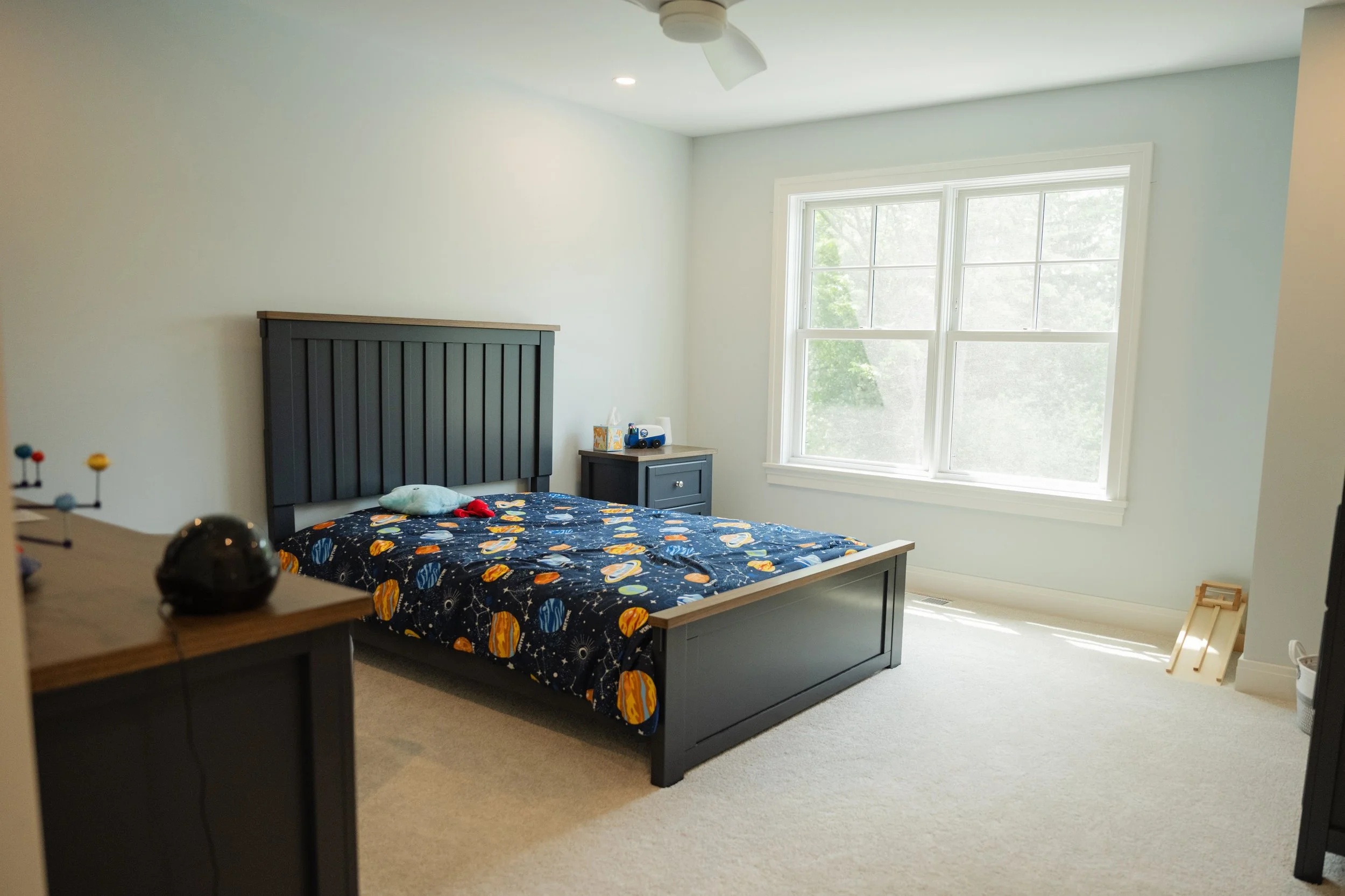 A bedroom with a large window allowing natural light. There is a dark-colored bed with a space-themed bedspread, a small nightstand with toys and tissues, and a ceiling fan. The walls are painted white, and the floor has a light-colored carpet.