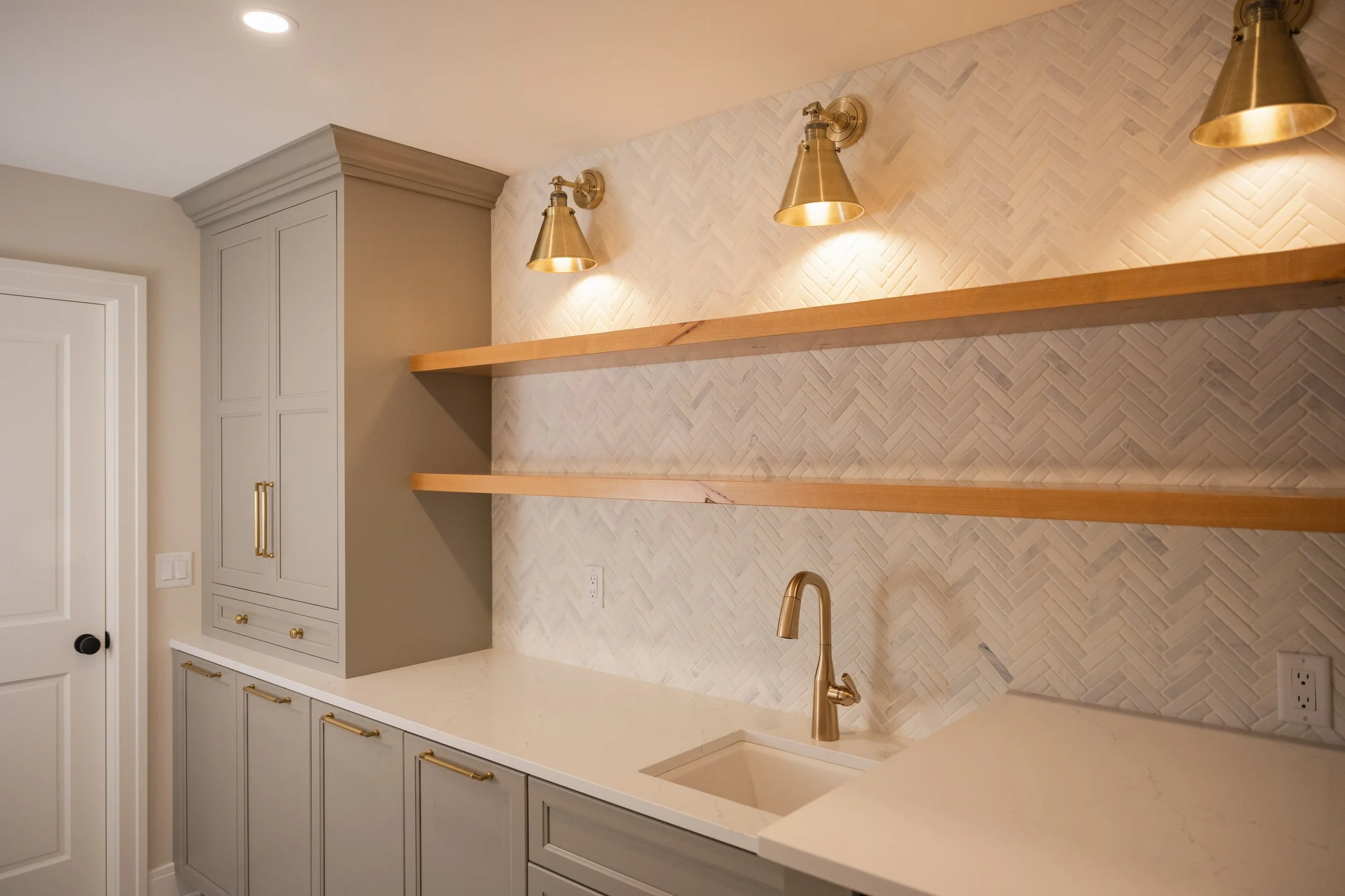 Kitchen with gray cabinets, white marble countertop, gold handles, gold fixtures, a herringbone tile backsplash, two wooden shelves, gold wall sconces, and a white door.