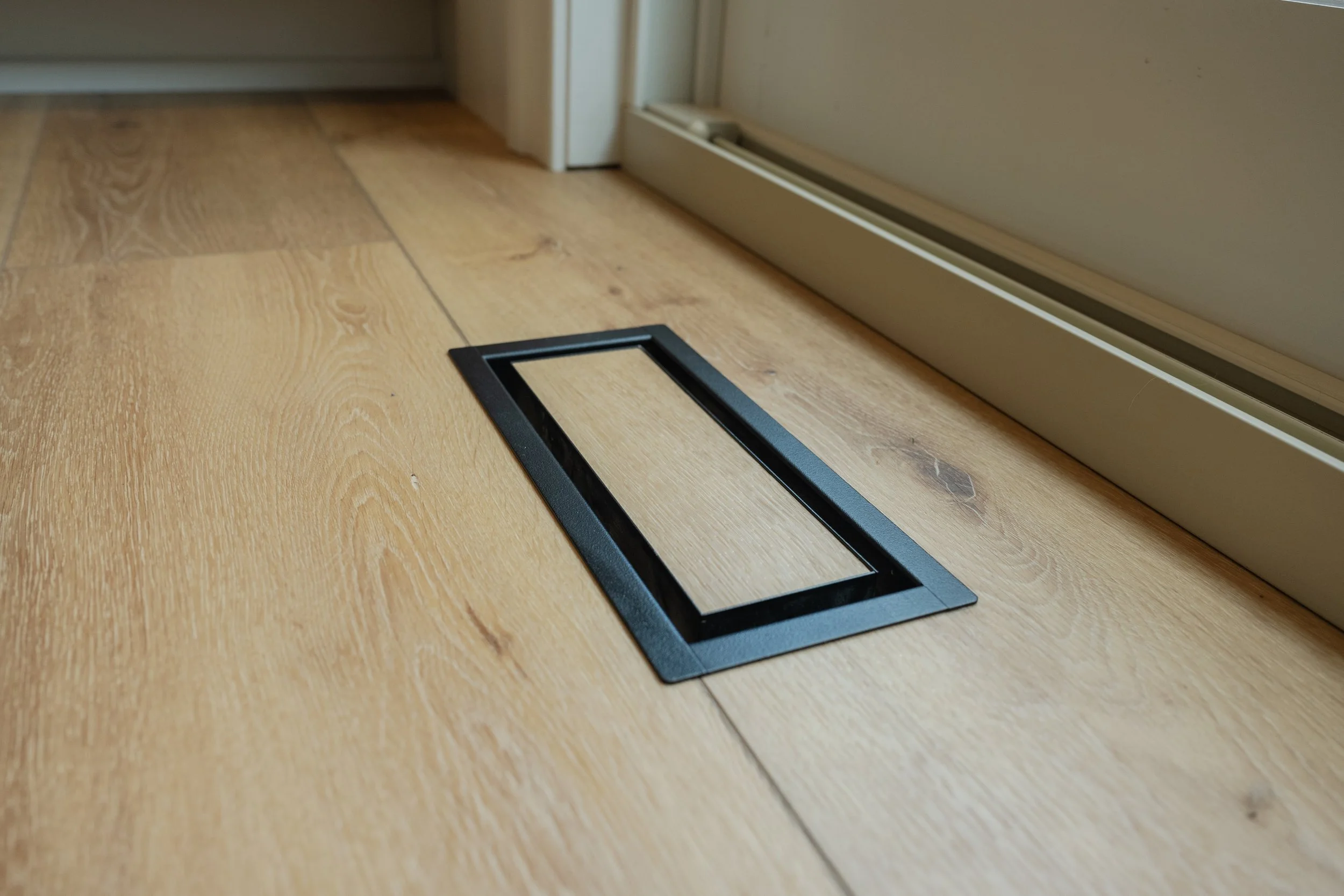 Close-up of a wooden floor with a rectangular black vent cover and a white baseboard heater along the wall.