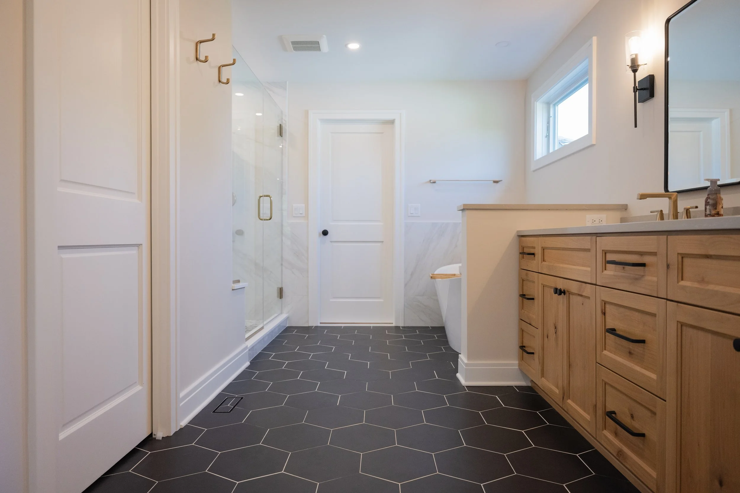A modern bathroom with black hexagonal floor tiles, a wooden vanity with black handles, a large wall mirror, wall-mounted light fixtures, a window, a glass shower with a gold handle, and a white door.