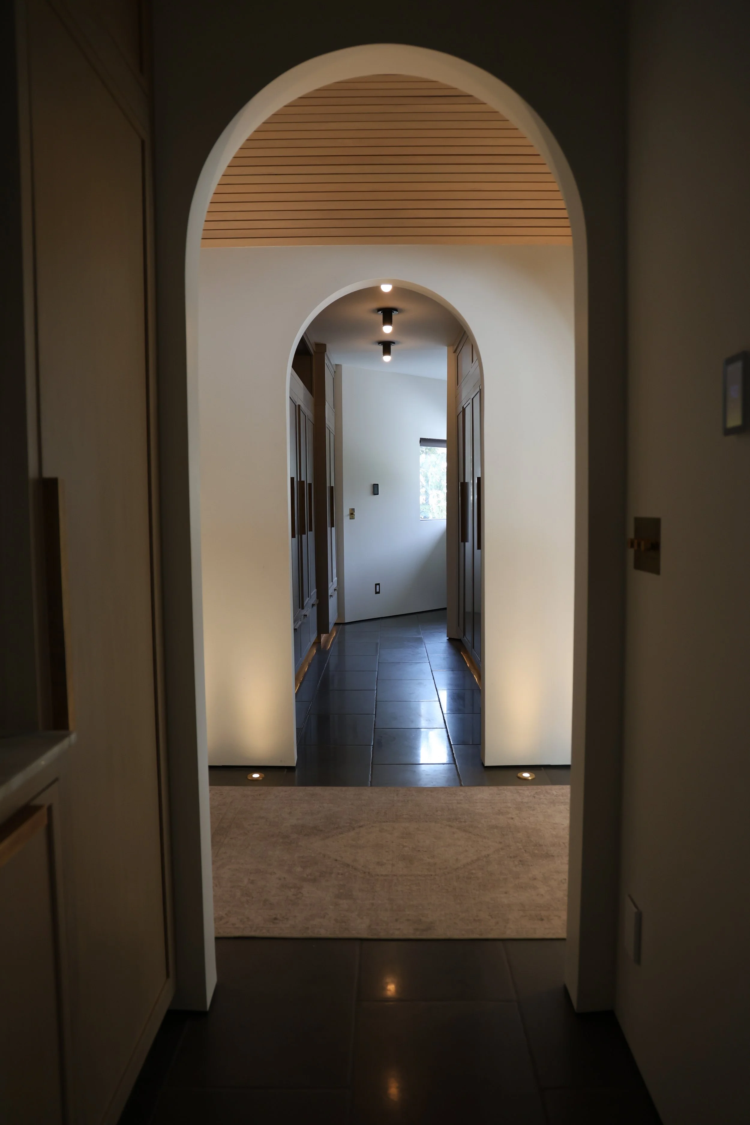 A hallway with dark flooring, light-colored walls, and wooden cabinetry, seen through an arched doorway with ceiling lights, a window at the end letting in natural light, and a beige rug.