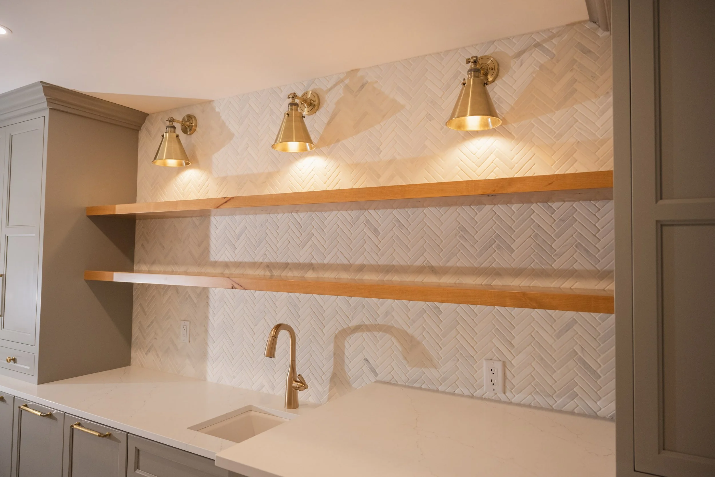 Modern kitchen with white cabinetry, a white countertop, beige herringbone tile backsplash, wooden shelves, gold fixtures, and a gold faucet.