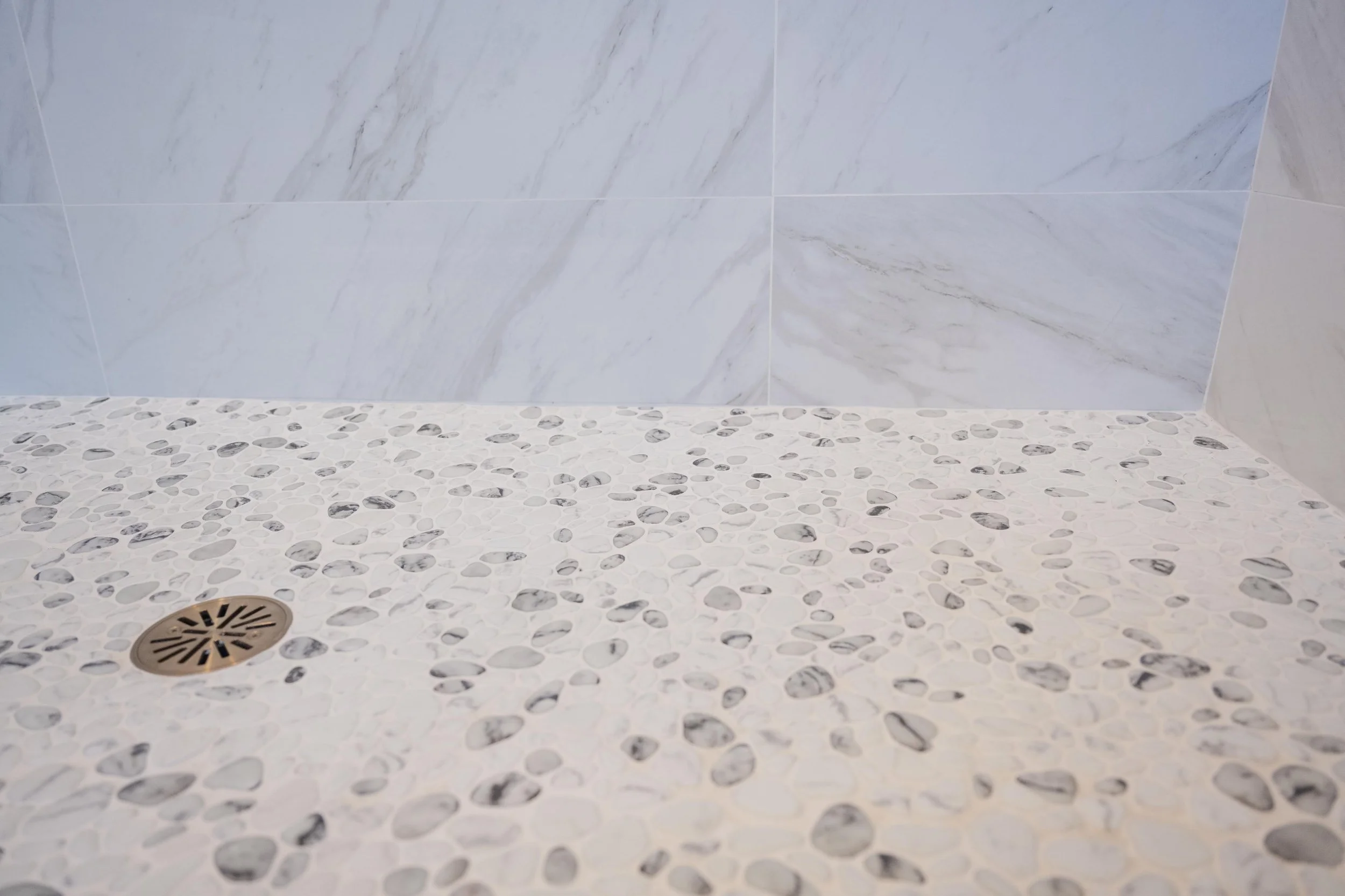 A bathroom floor with white pebble tiles, a circular metal drain, and a wall of large white marble tiles with gray veining.