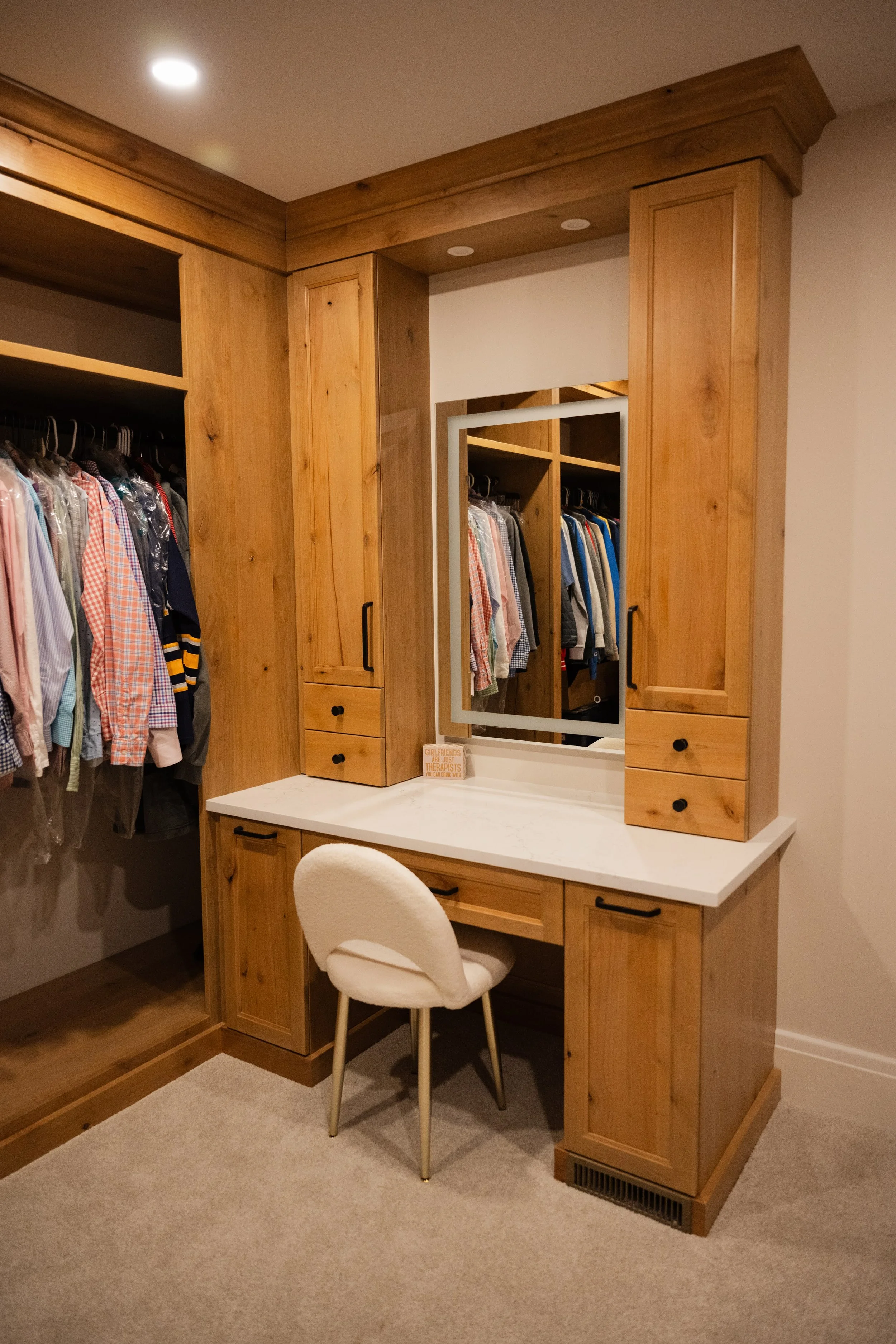 Wooden closet with a built-in vanity, mirror, drawers, and shelves, situated in a bedroom with a beige carpet and a white chair in front.
