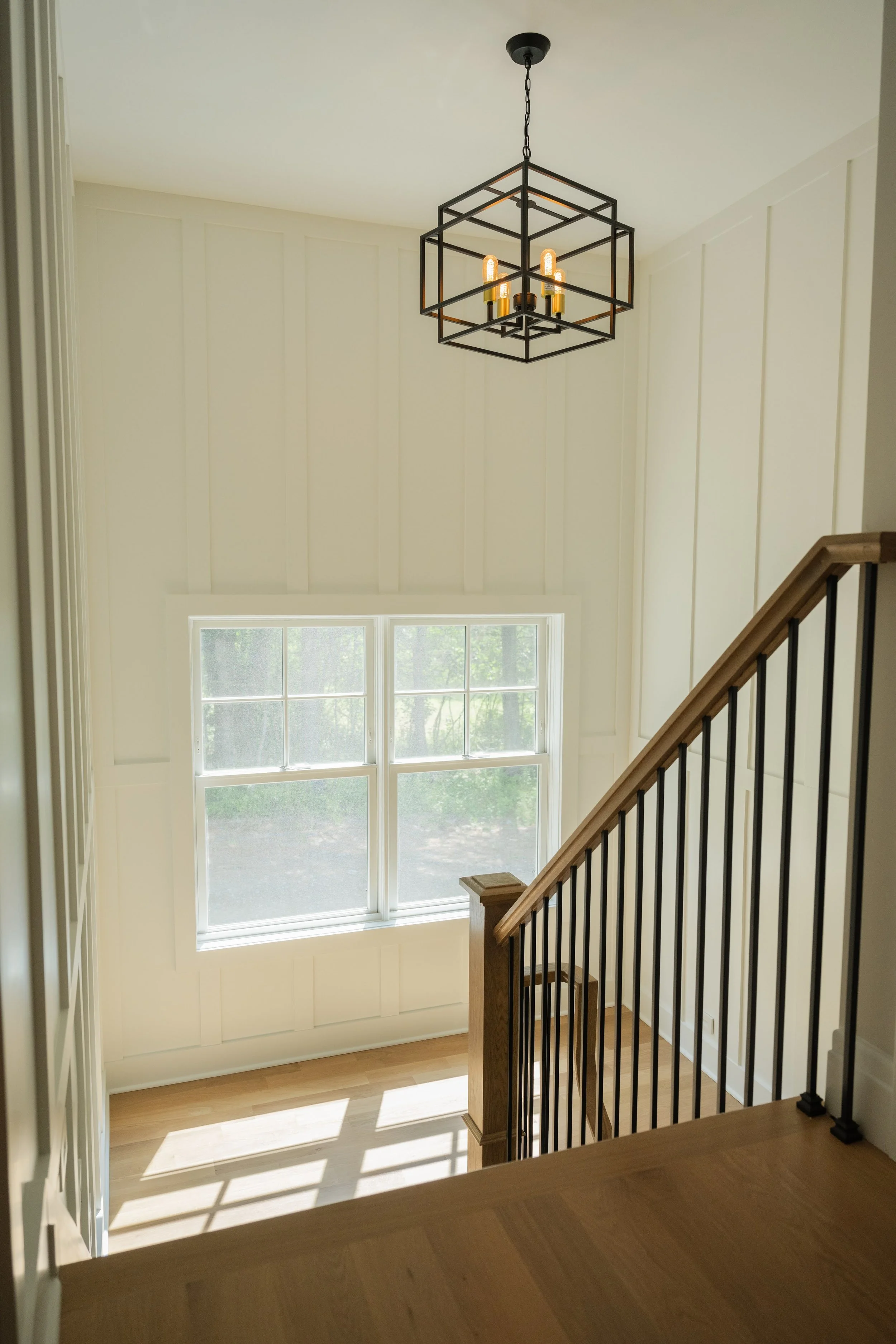 Interior view of a staircase landing with a large window, white painted wood panels, and a modern geometric black metal chandelier hanging from the ceiling.