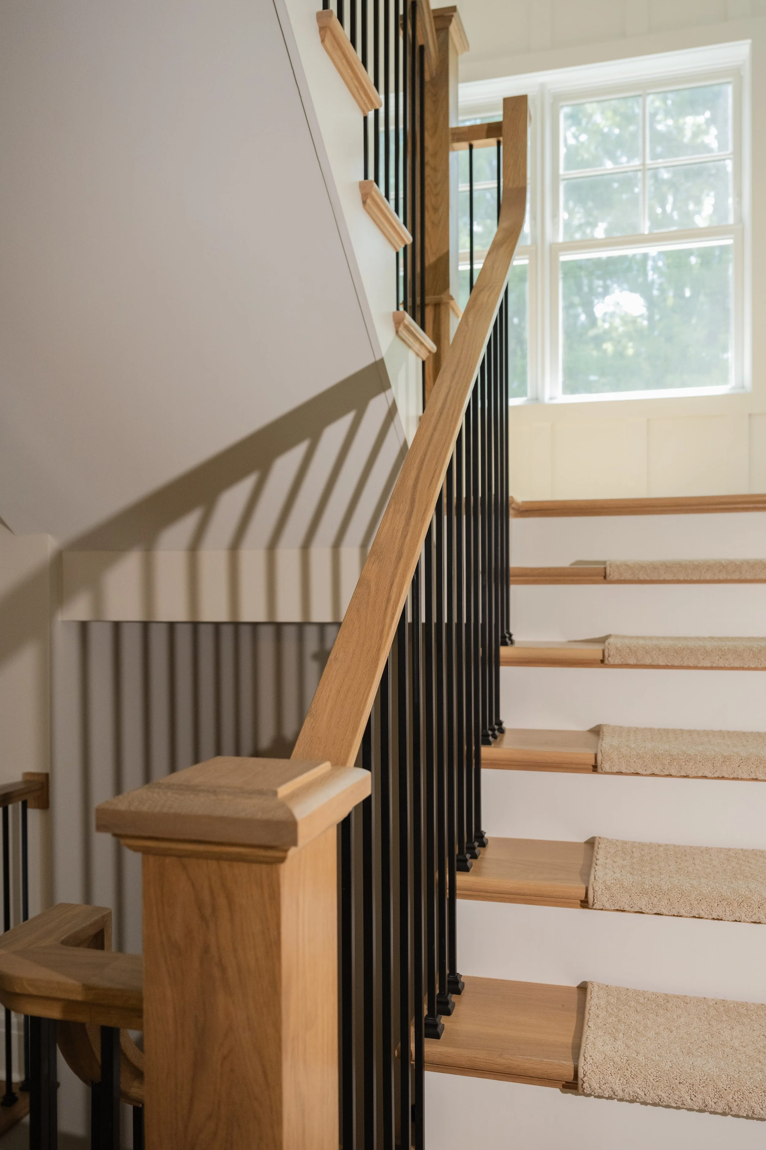 A staircase with wooden handrails and beige carpeted stairs, black metal balusters, and a large window in the background letting in natural light.