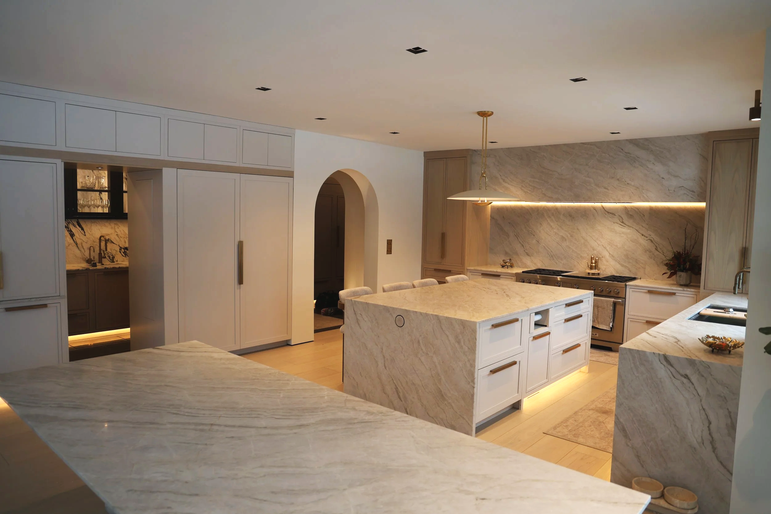Modern kitchen with white marble countertops, a large island, built-in appliances, and wooden cabinetry, with a hanging light fixture and a marble backsplash.