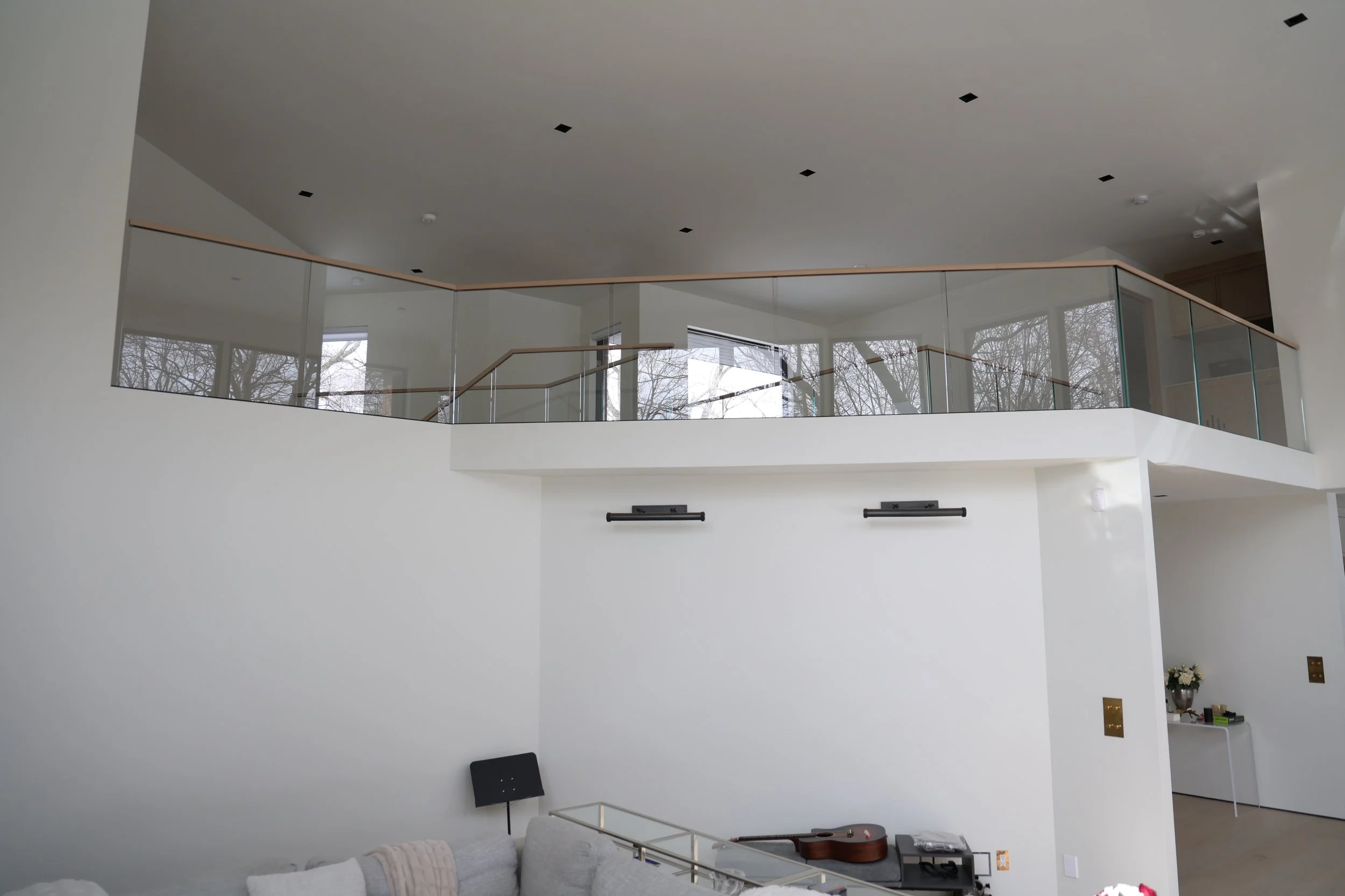 Interior view of a modern, spacious house with high ceiling, large windows, and a mezzanine with glass railing. There is a living area with white furniture, a guitar on a table, and decorative plants.