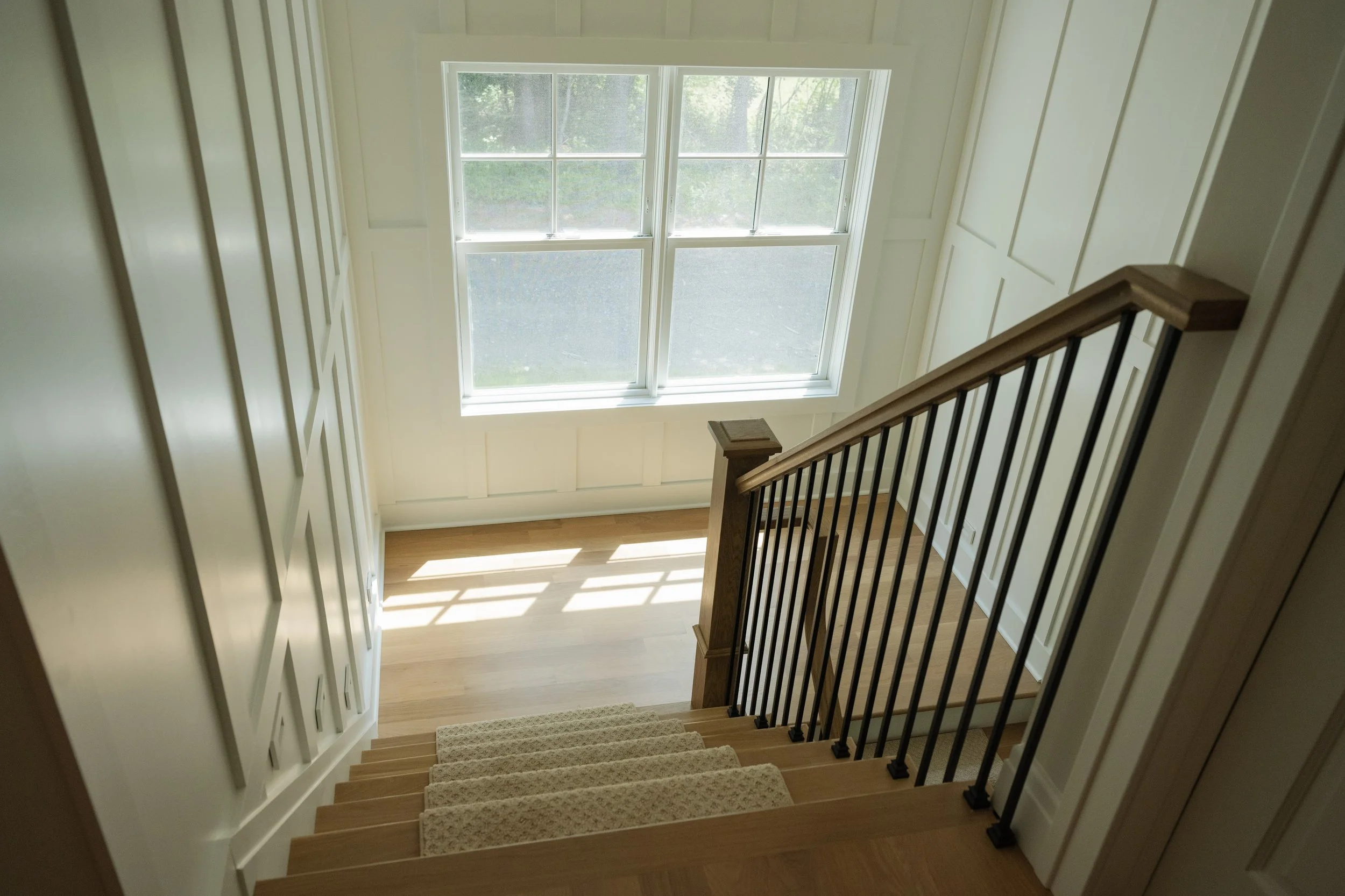 Interior view of a staircase with wooden steps, black railing, and a large window at the top letting in natural light.