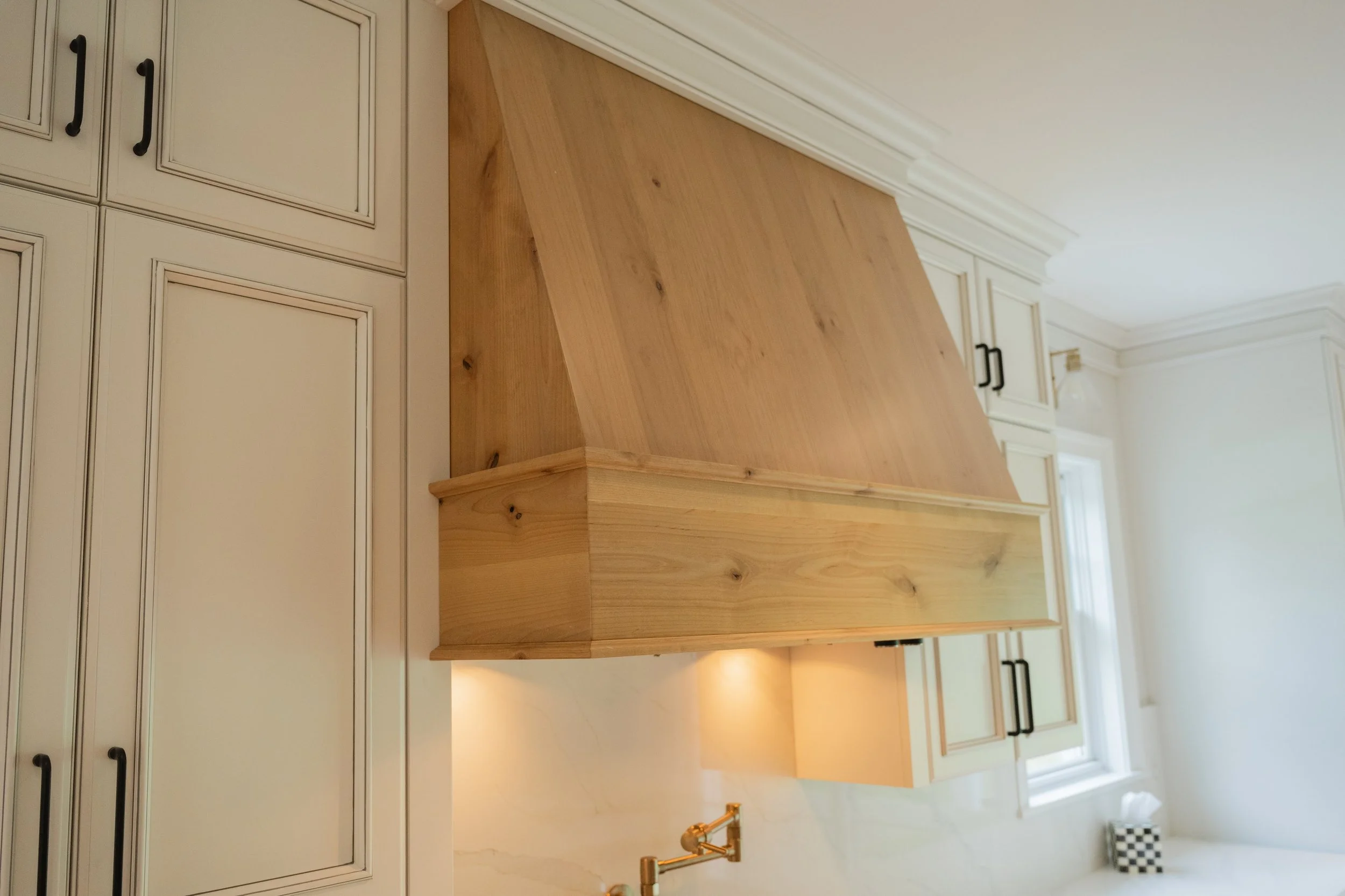 Custom wooden stove hood, custom cabinetry with black handles, a wooden kitchen island, a double sink with a brass faucet, and windows with white blinds.