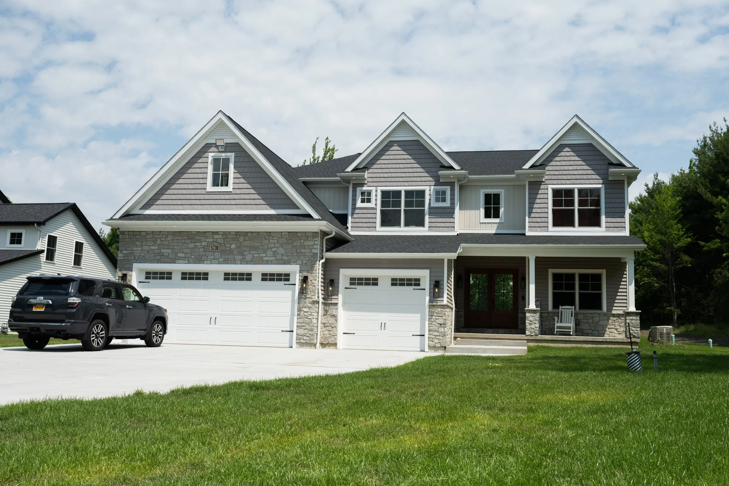 Front view of a two-story suburban house with a gray and white exterior, three garage doors, a black SUV parked in the driveway, and a green lawn in the foreground under a partly cloudy sky.