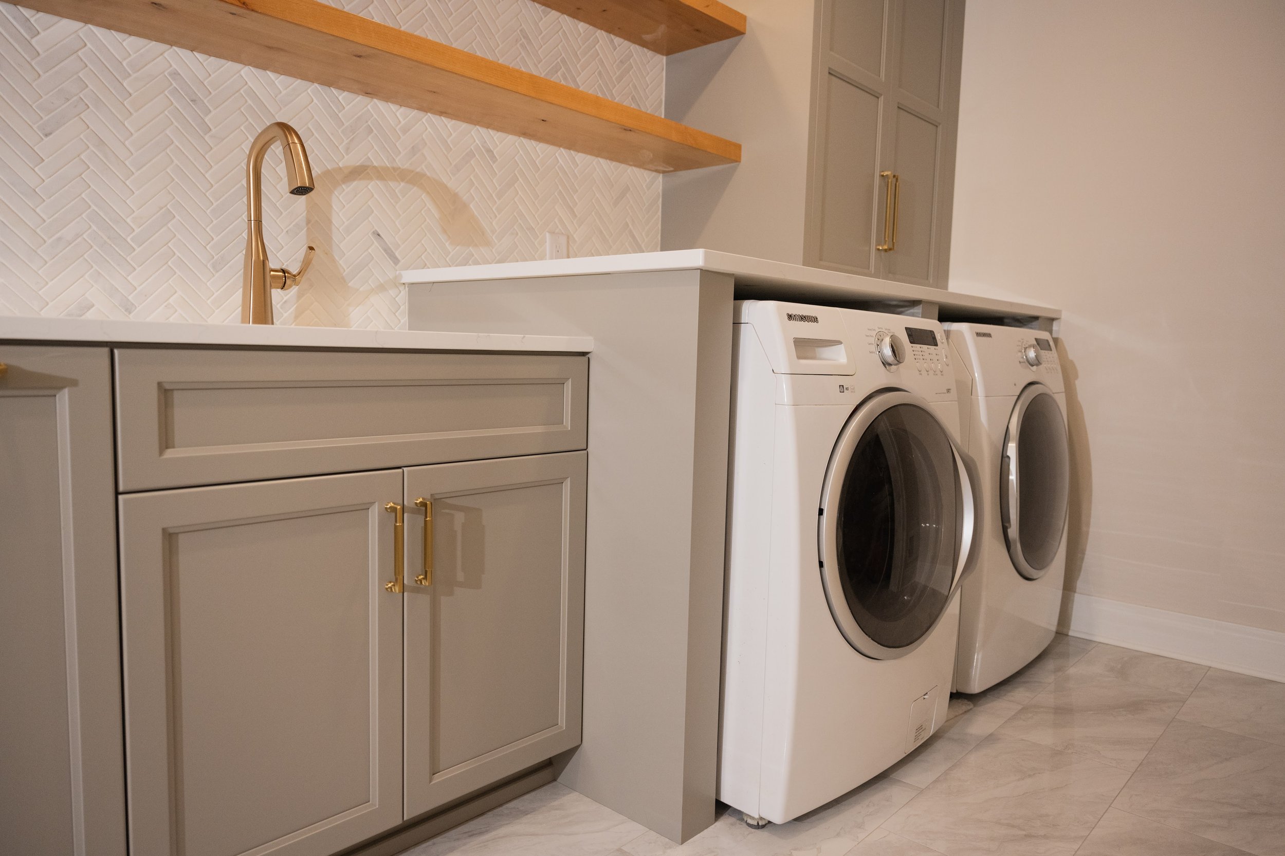 Laundry room with gray cabinets, a white countertop, a gold faucet over the sink, open wooden shelves, and two front-loading washing machines.