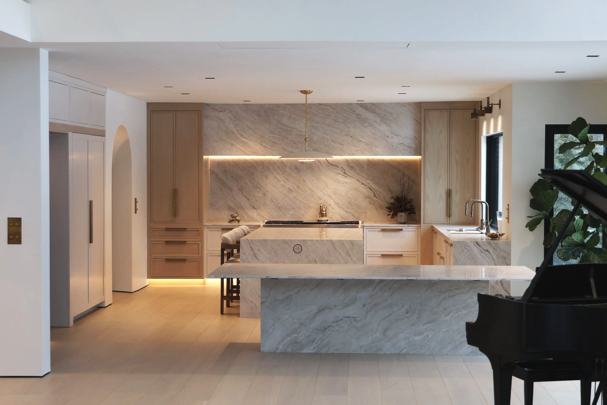 Modern kitchen with marble countertops and backsplash, wooden cabinetry, a black grand piano, and a window with greenery outside.