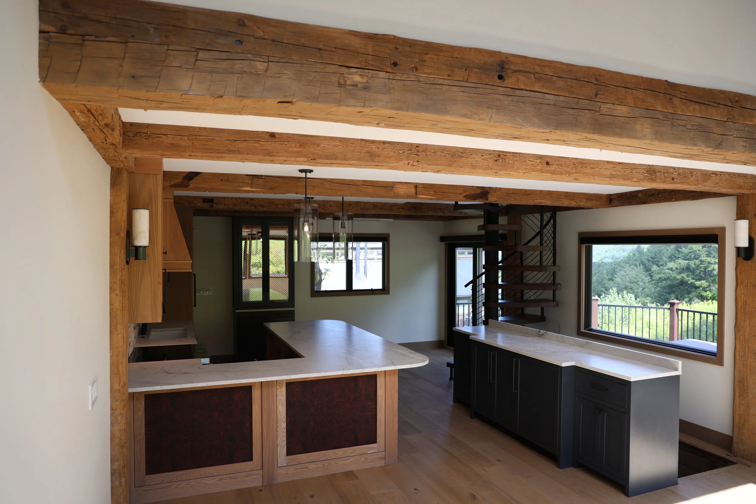 A kitchen with wooden beams on the ceiling, a marble-topped island, black cabinets, large windows, and a spiral staircase.