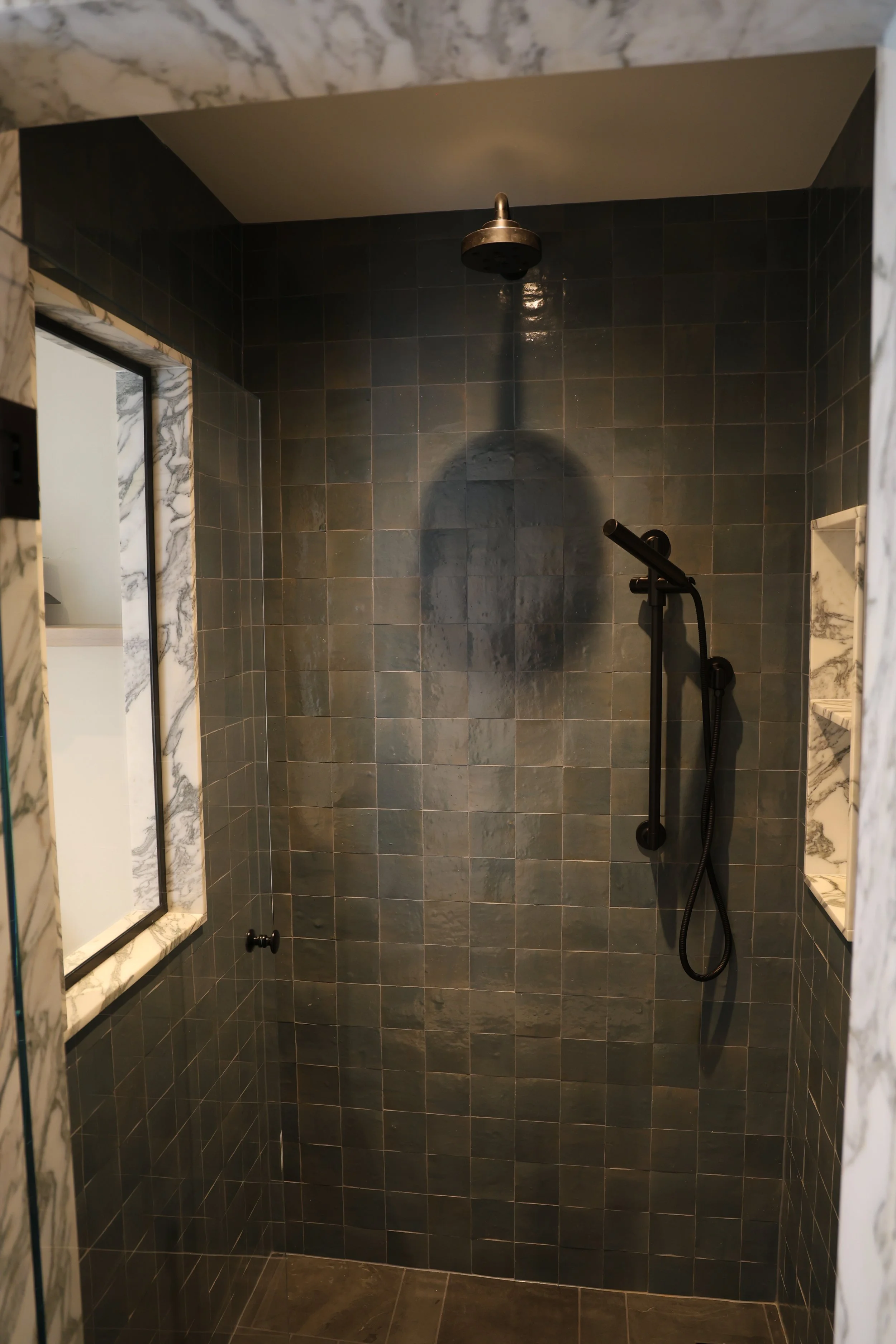 A modern shower stall with dark gray tiles on the walls, a ceiling-mounted shower head, and a handheld shower wand. The frame of a mirror is visible on the left, and marble accents are seen along the edges of the doorway.