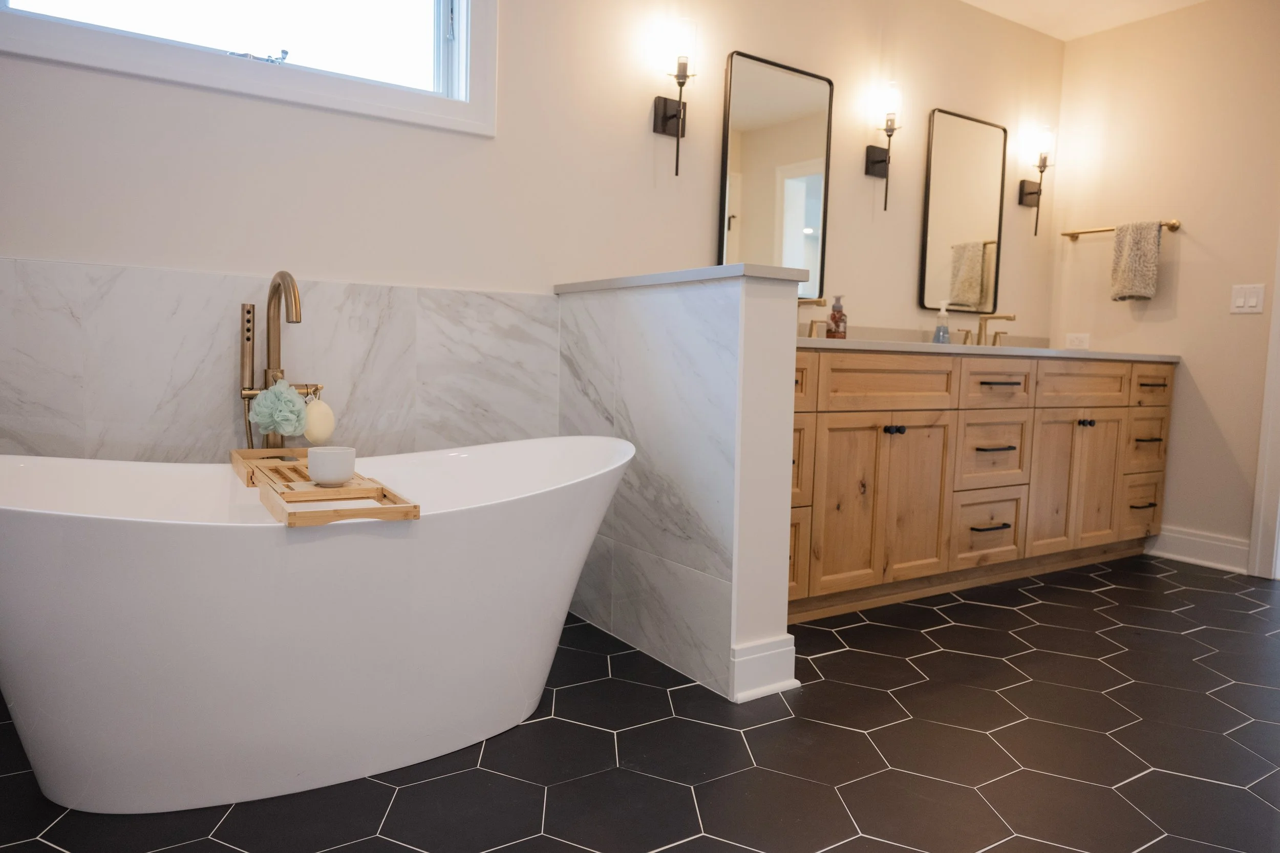 Modern bathroom with a white freestanding bathtub, black hexagon floor tiles, a wooden vanity with black hardware, a large mirror, and black wall-mounted light fixtures.
