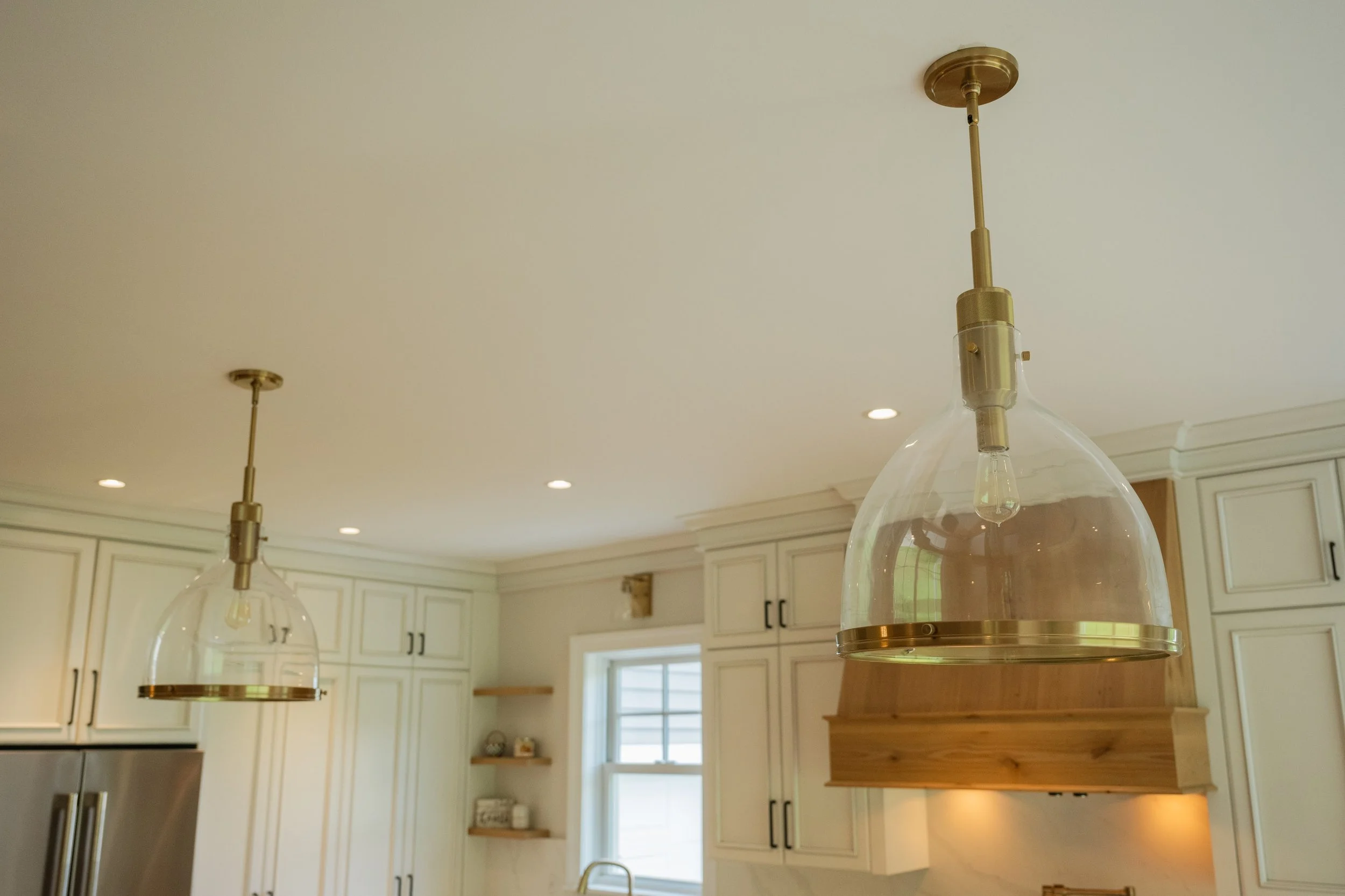 Two modern pendant light fixtures hanging from a white ceiling in a kitchen with white cabinets and a window.