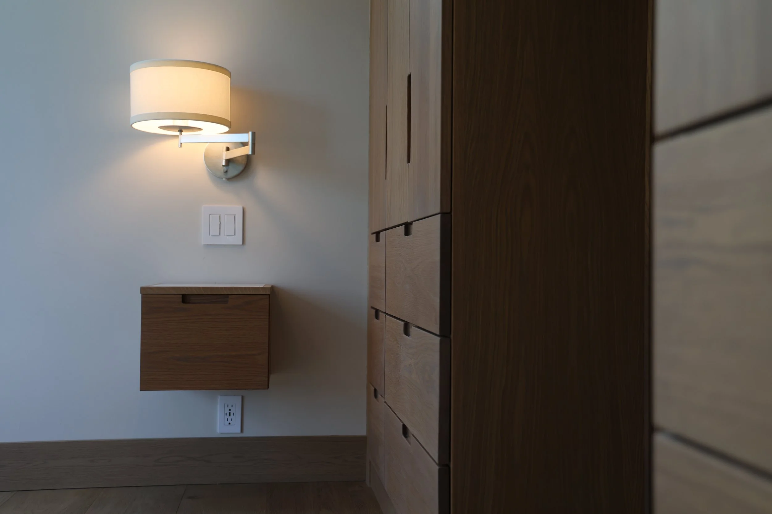 A wall-mounted lamp with a white shade, a white electrical switch, and a power outlet on a white wall, with a wooden cabinet and drawers below.