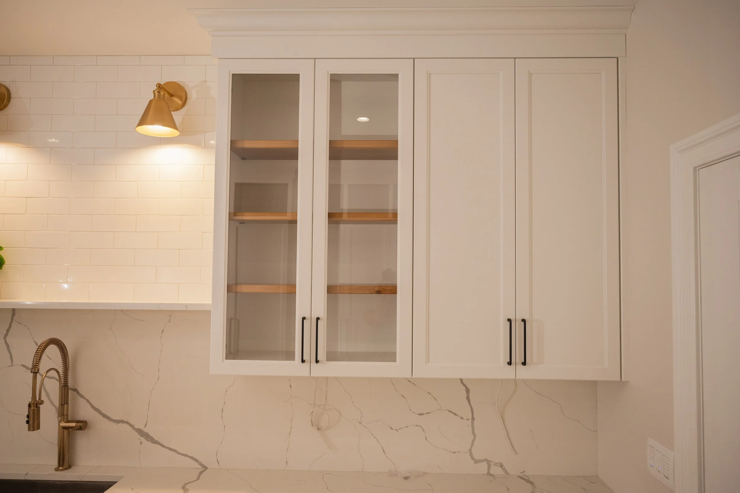 Kitchen with white cabinetry with black handles, glass doors, and open shelves, white subway tile backsplash, beige marble countertops and flooring, gold wall sconce lights, brass faucet, and a white ceiling with crown molding.