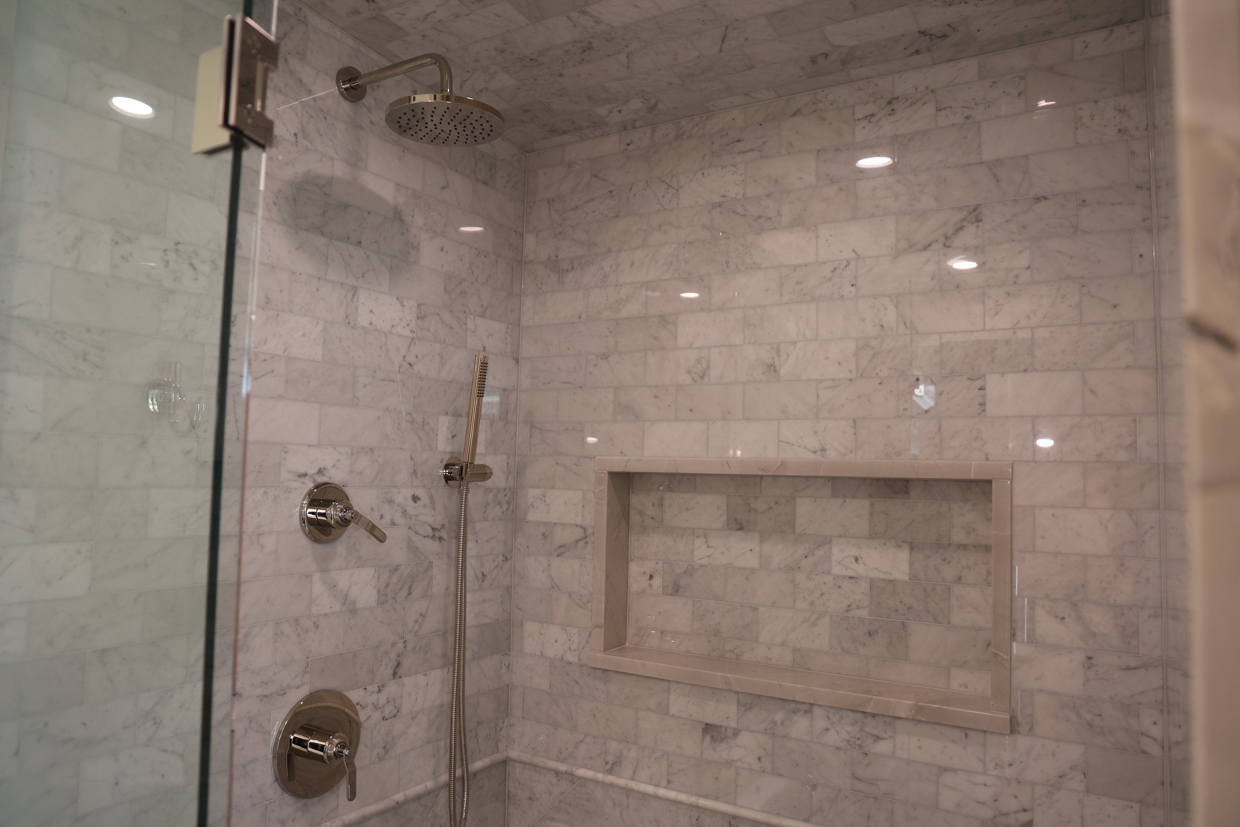 A marble shower area with a rain showerhead, a handheld shower, and a built-in marble shelf, enclosed by a glass door.