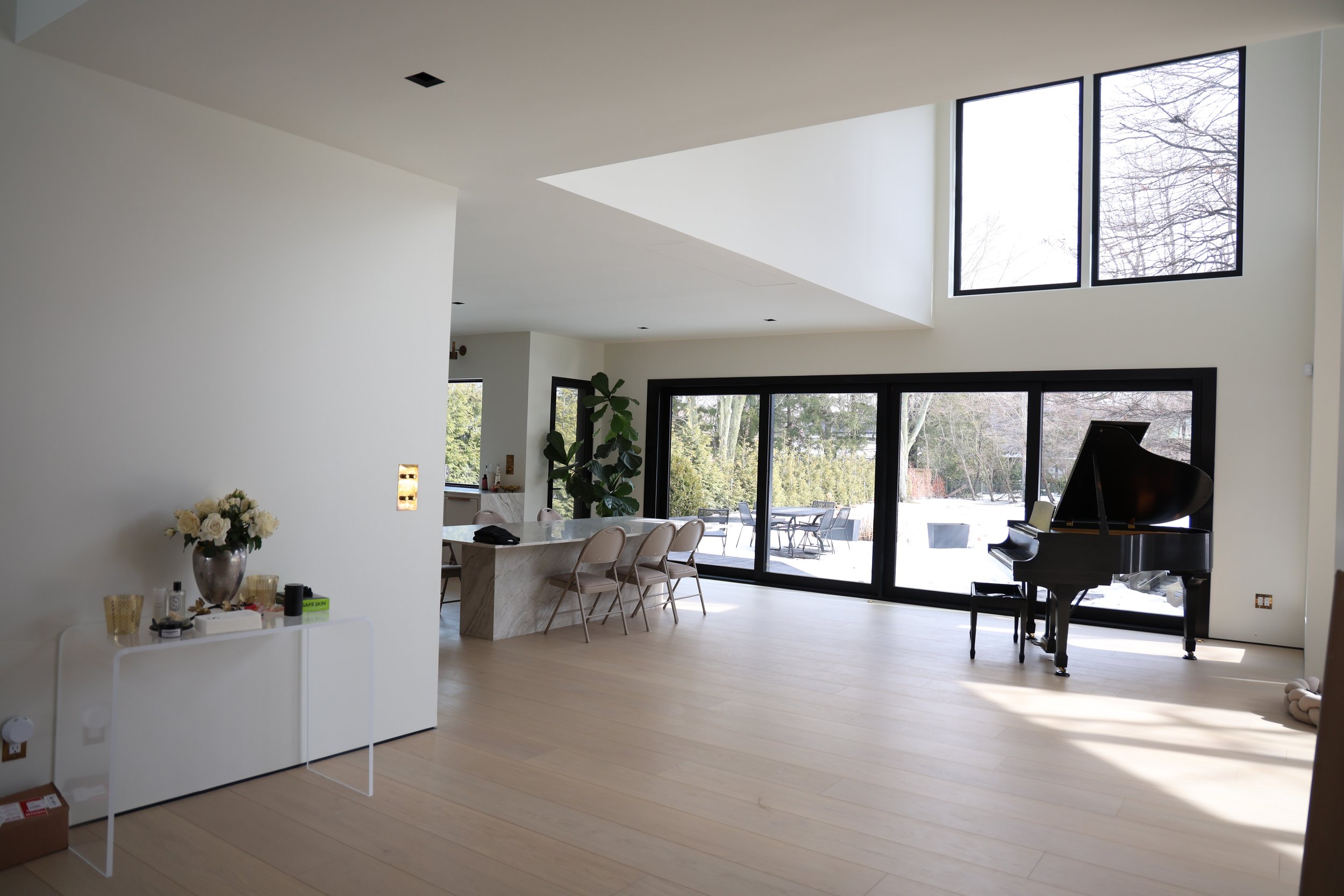 Modern living room with high ceiling, large black-framed windows, and a grand piano. A dining table with beige chairs is visible, along with a vase of white roses and a small white table near the wall. The room has light-colored wooden flooring and g