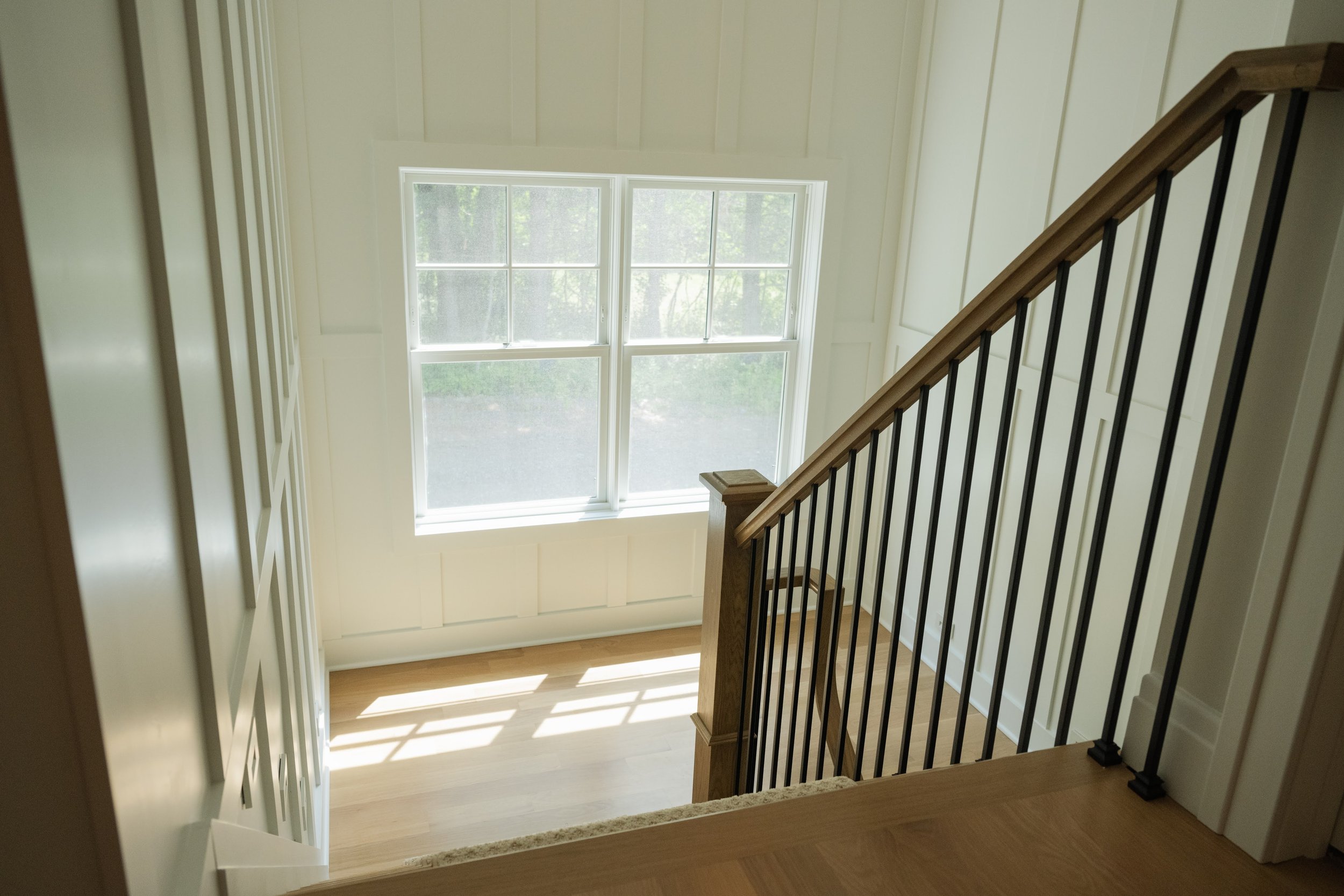 View of a staircase with a wooden handrail and black balusters leading down to a bright room with large window letting in natural light.
