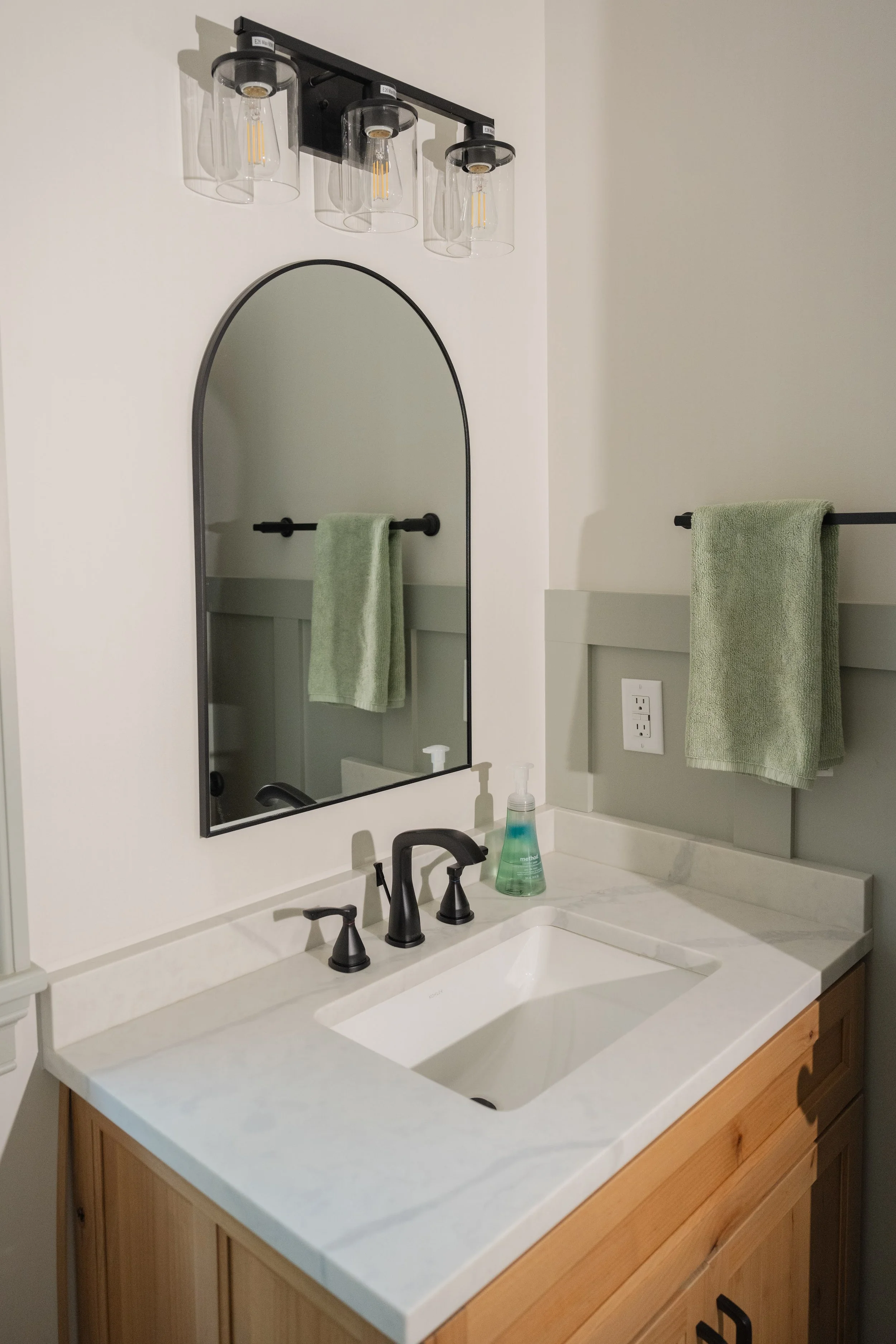 Bathroom vanity with a white marble countertop, black fixtures, a styled black-framed mirror, two green towels on black towel rods, a bottle of hand soap, and a three-light fixture with clear glass shades above.