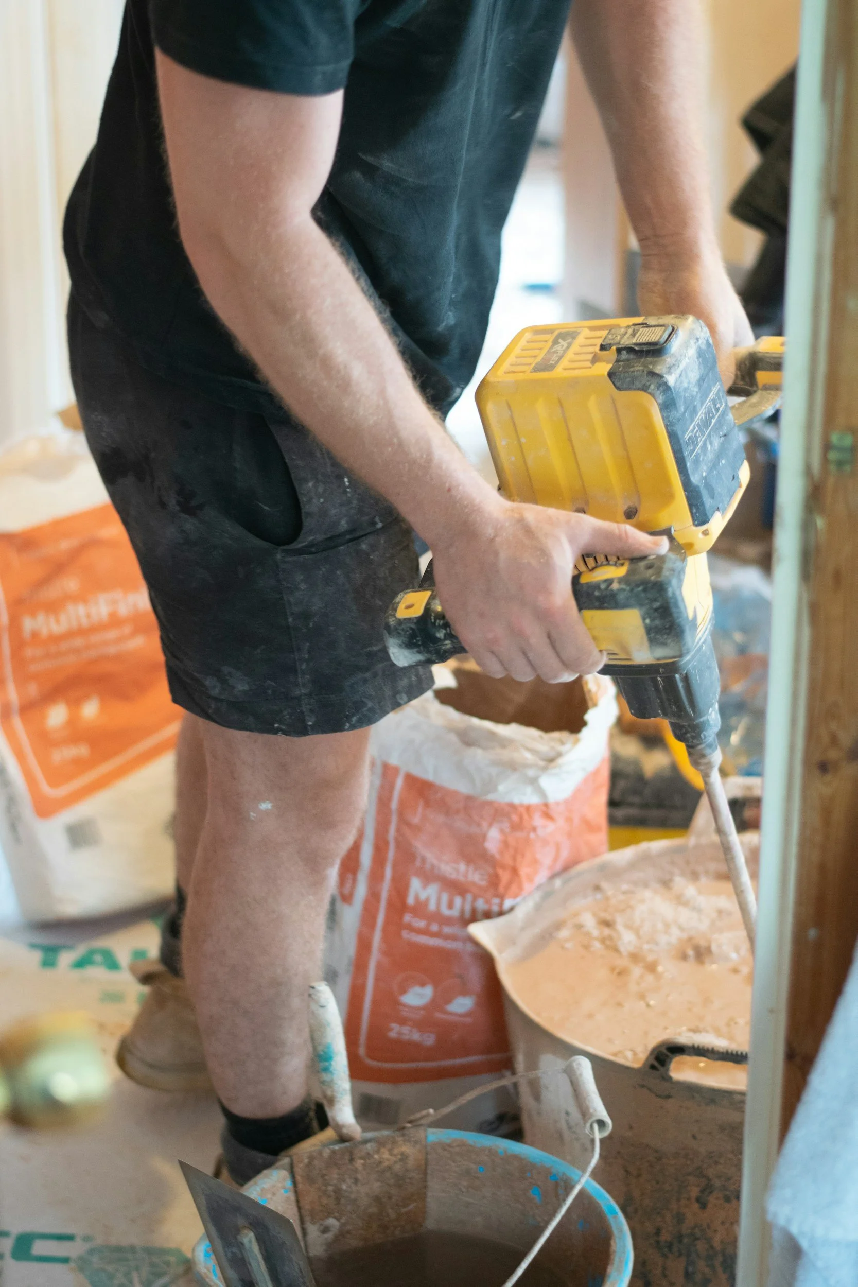Person arbeitet mit einem elektrischen Werkzeug bei Bauarbeiten. Im Hintergrund sind Baumschüttgut und Baumaterialien sichtbar.