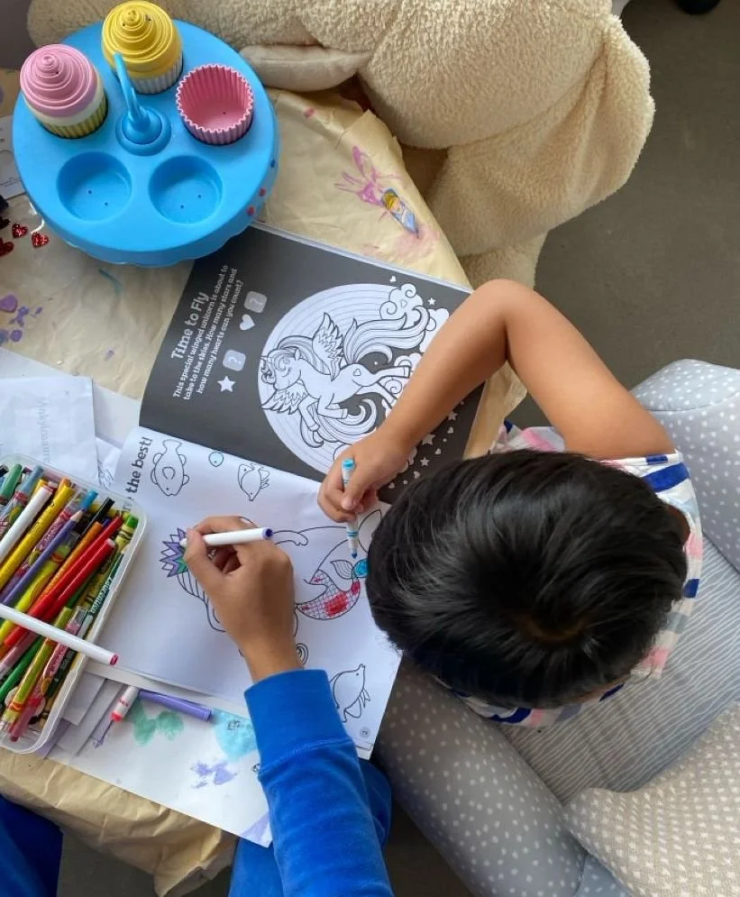 A child coloring a unicorn illustration in a coloring book, with markers on a table, and a cupcake decorating toy set nearby.
