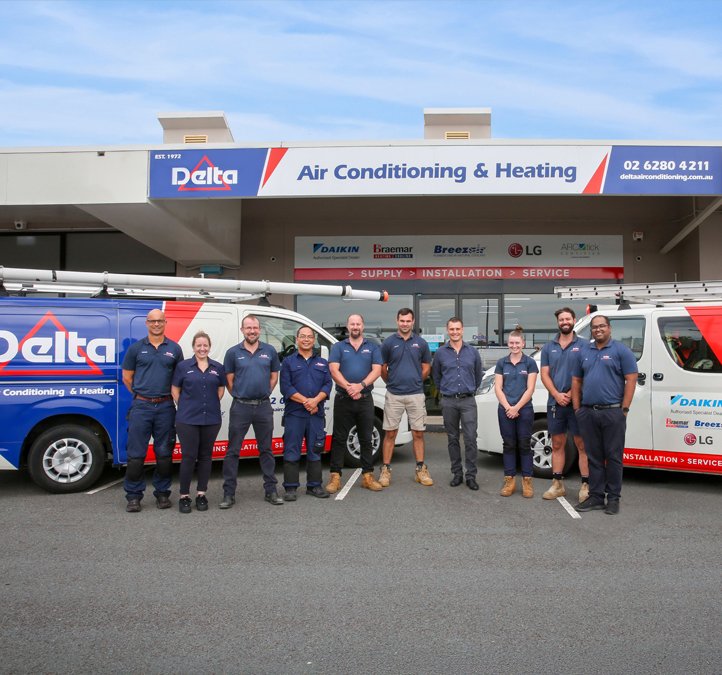 Group of nine HVAC technicians standing in front of two service vans outside an air conditioning and heating business storefront.
