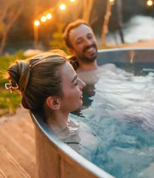 A man and woman relaxing in an outdoor hot tub at sunset with warm lighting and trees in the background.