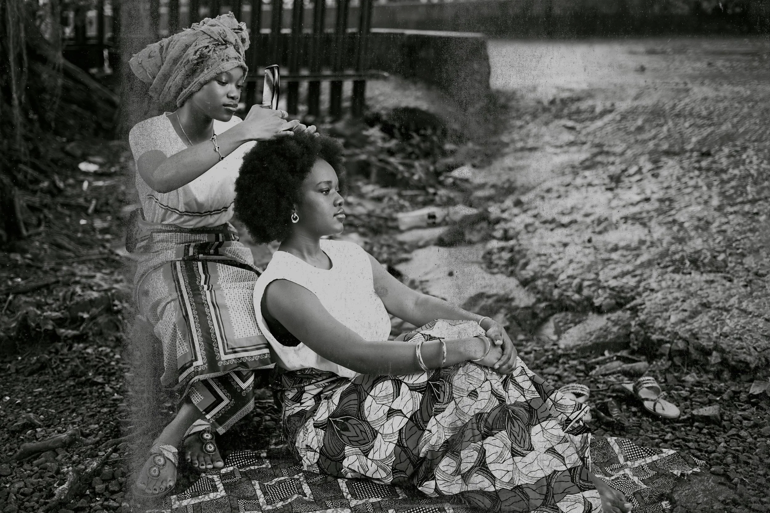 Two women, one sitting on the ground and the other standing, on a rocky outdoor surface with a fence in the background. The standing woman is holding a mirror and appears to be adjusting the seated woman's hair, who is wearing patterned skirt and jewelry.