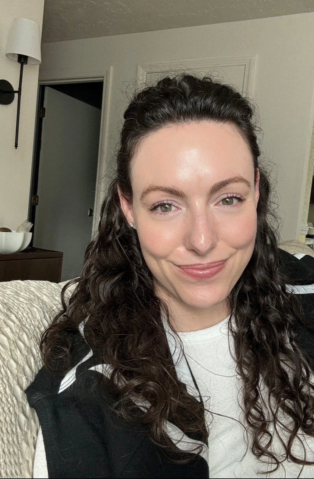 A woman with curly dark hair and light skin smiling indoors, wearing a white shirt and black cardigan.