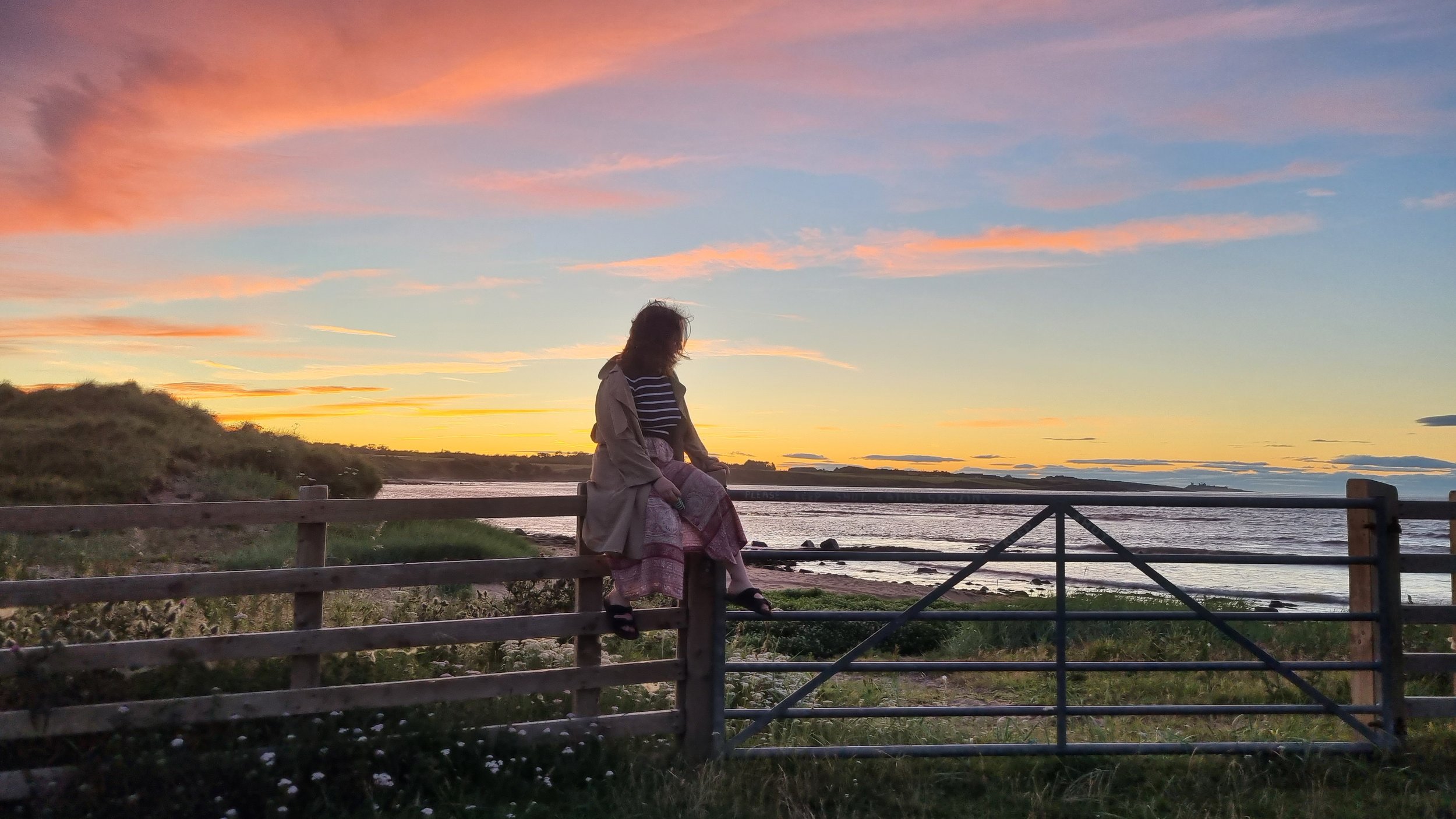 A woman sitting on a wooden fence near a body of water, watching a colorful sunset sky with pink, orange, and purple clouds.