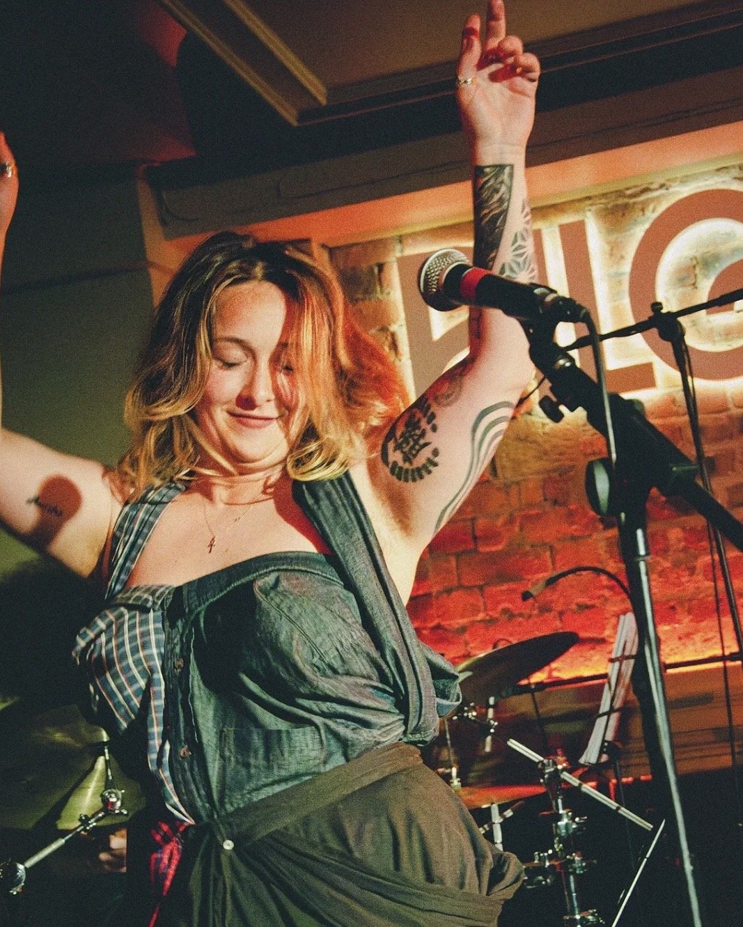 A woman with tattoos and blonde hair dancing on stage with her eyes closed, raising one arm, in front of a microphone with a drum set and brick wall background.