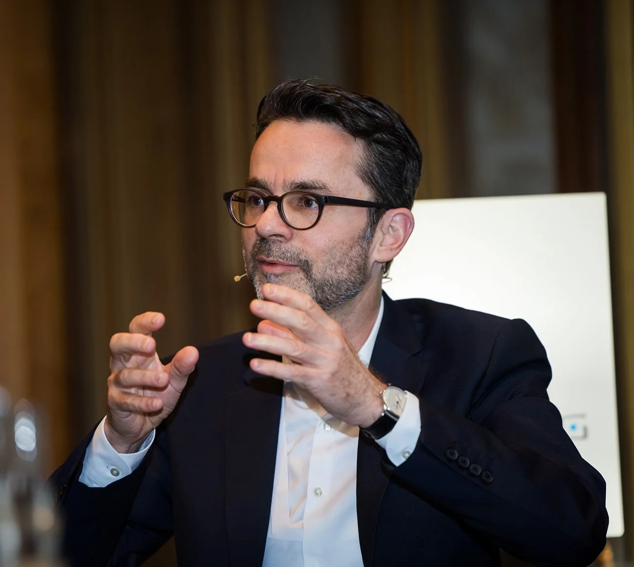 Guido Schommer mit Brille in einem Anzug spricht bei einer Veranstaltung, er trägt eine Uhr und eine kleine Mikrofonschlinge am Ohr.
