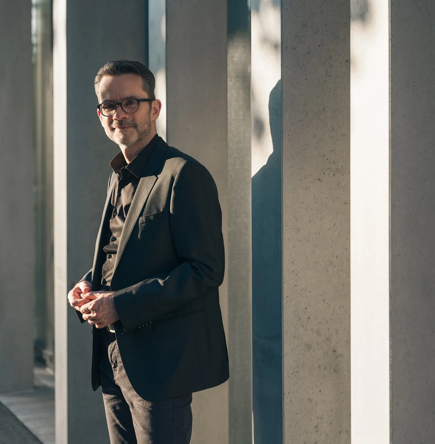Guido Schommer trägt einen schwarzen Anzug mit Hemd und Brille, steht neben einer Wand aus Betonpfeilern im Sonnenlicht.